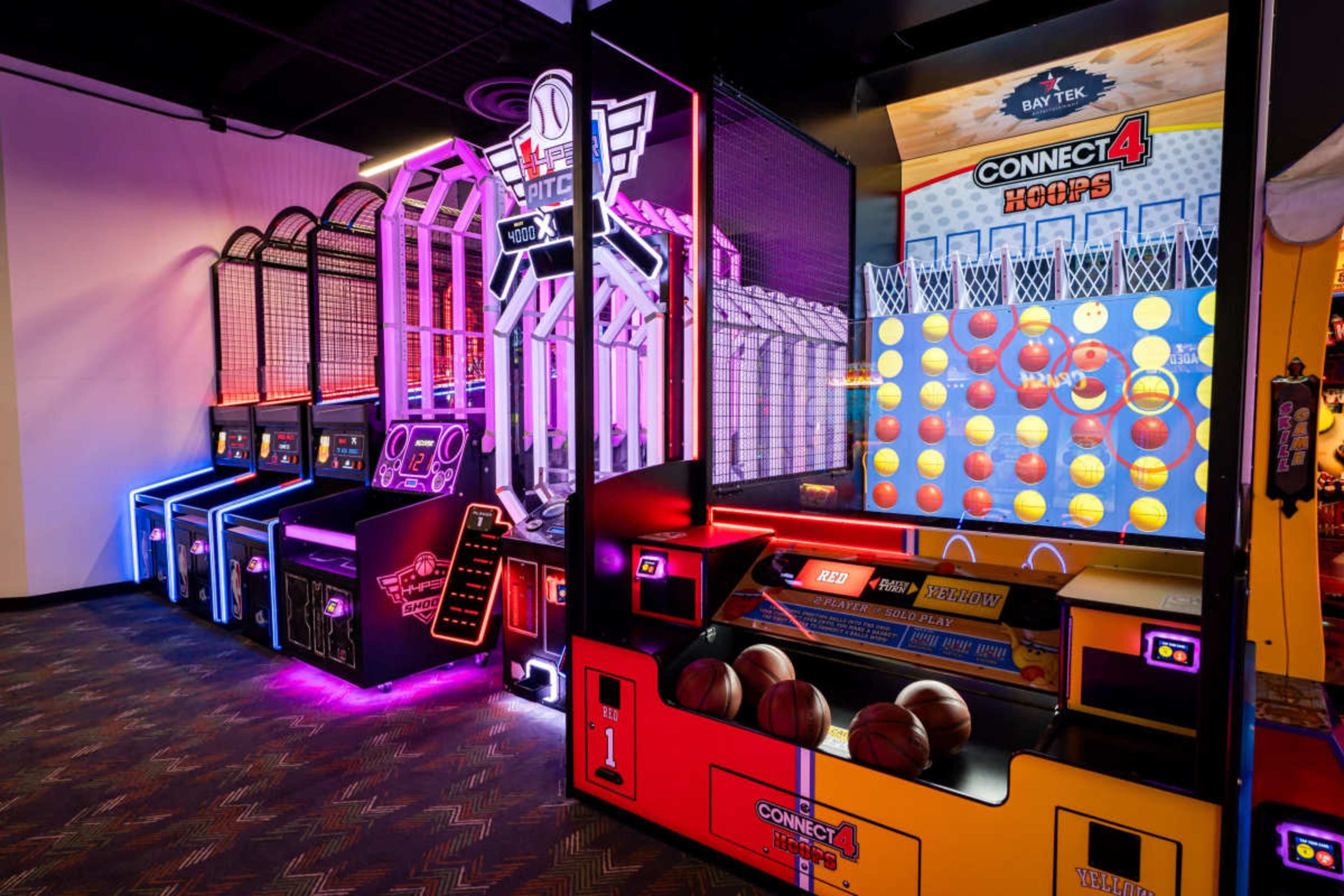 The image shows a brightly lit arcade featuring various games, including a basketball shooting game and a Connect 4 hoops game, set against a vibrant backdrop.