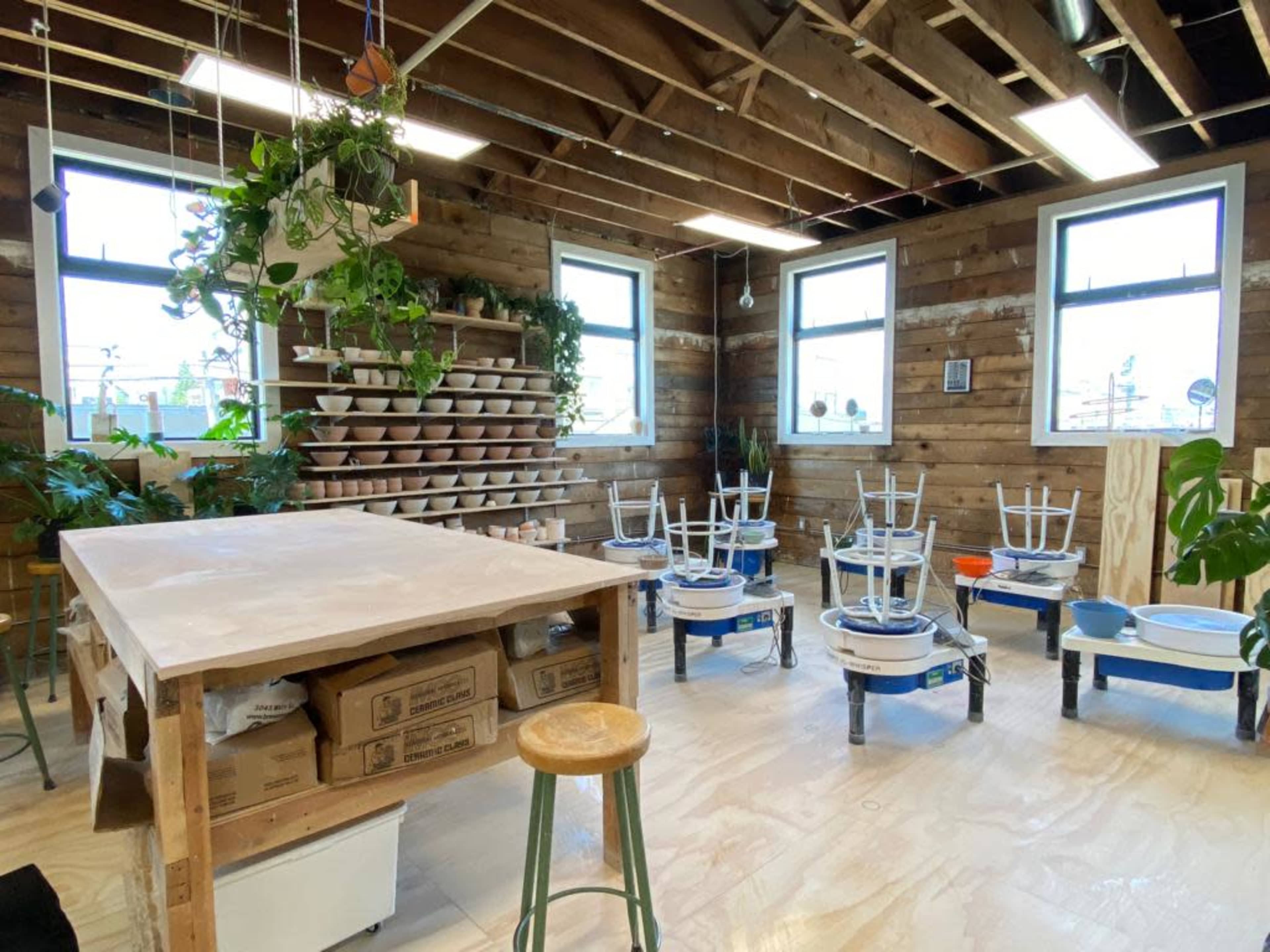 A pottery studio with wooden walls, a large work table, and several pottery wheels arranged in a circular layout, surrounded by plants and natural light from windows.