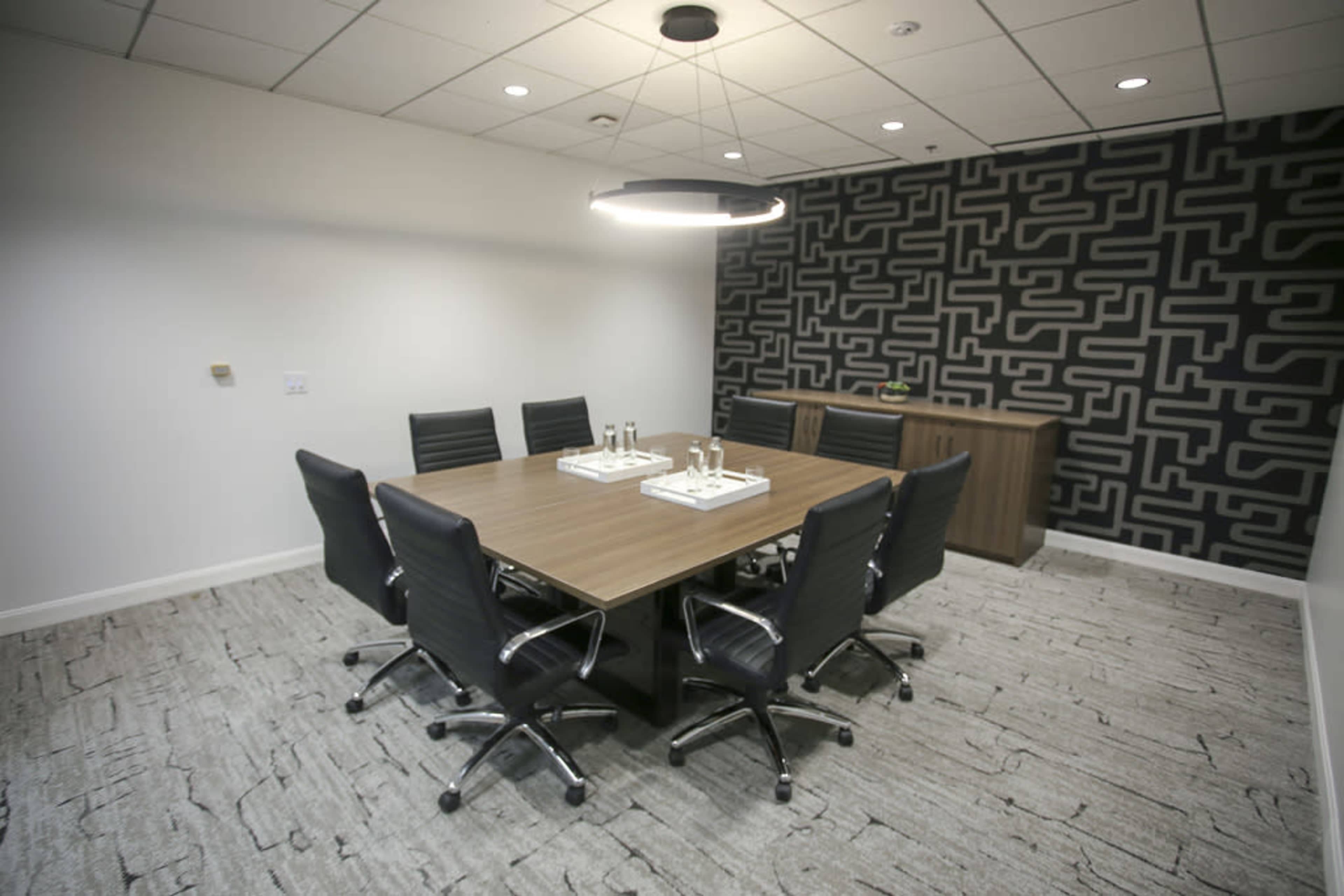 A conference room features a large wooden table surrounded by six black swivel chairs, with a patterned wall and modern overhead lighting.