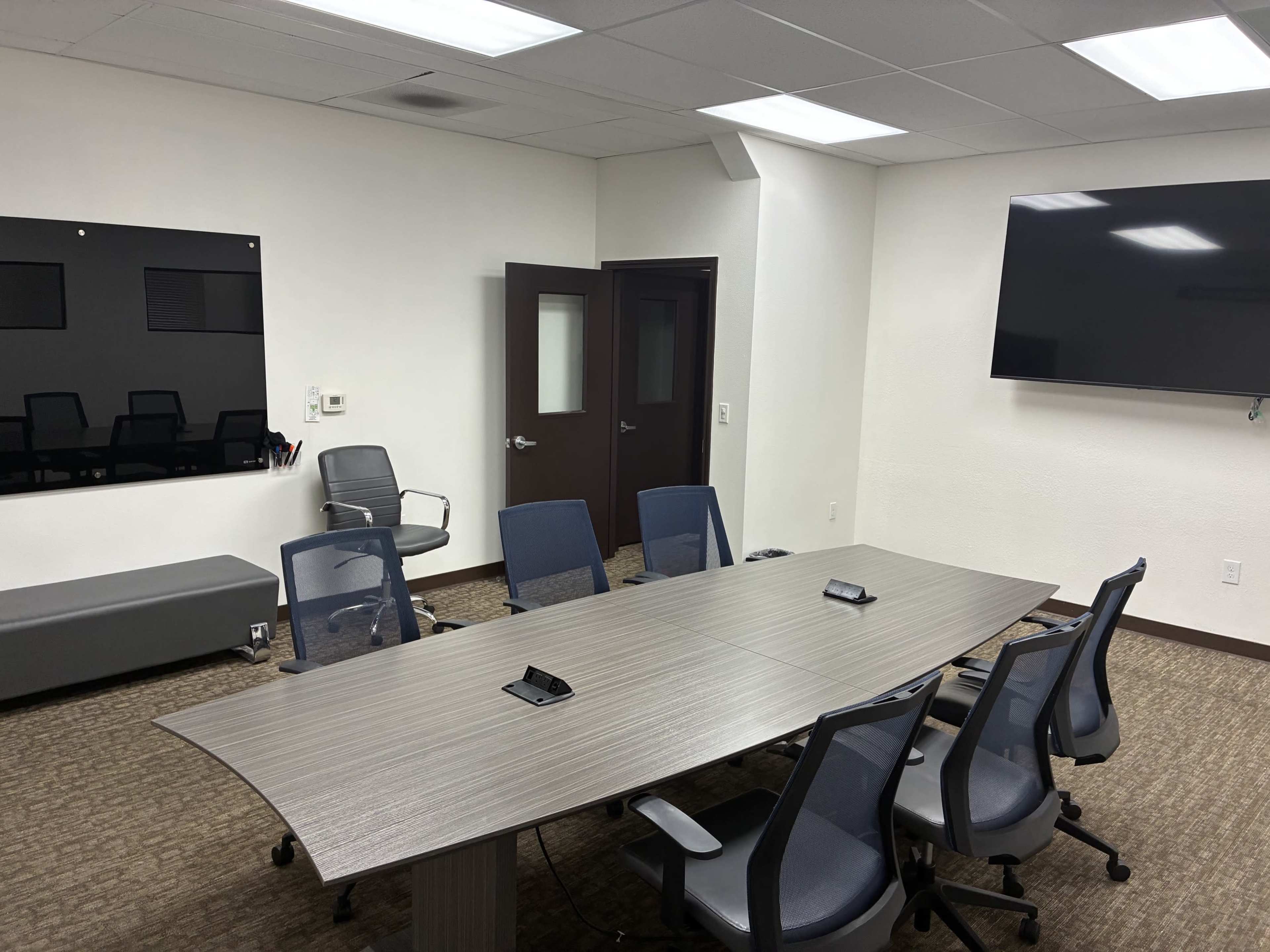 The image shows a modern conference room with a long table surrounded by six blue office chairs, a large television mounted on the wall, and a black panel for presentations.