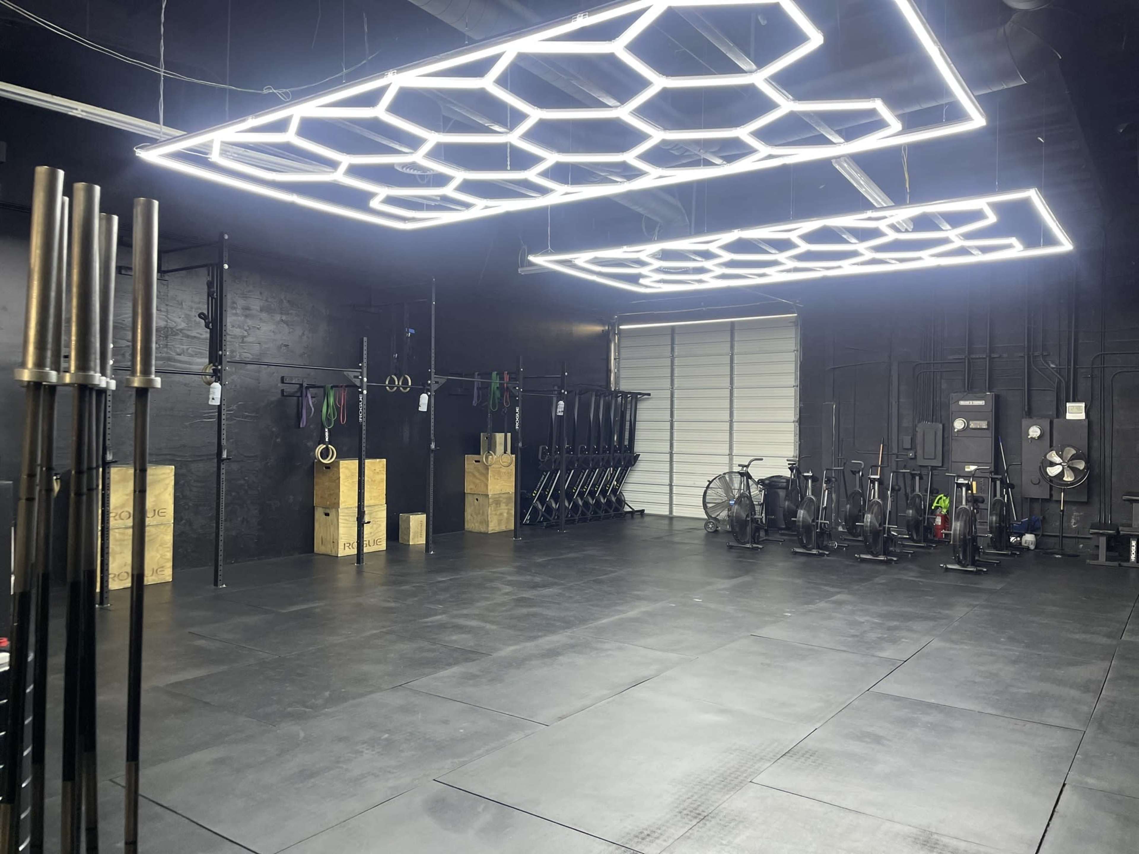 A spacious gym interior with hexagonal light fixtures, black walls, various exercise equipment, and a large overhead door.