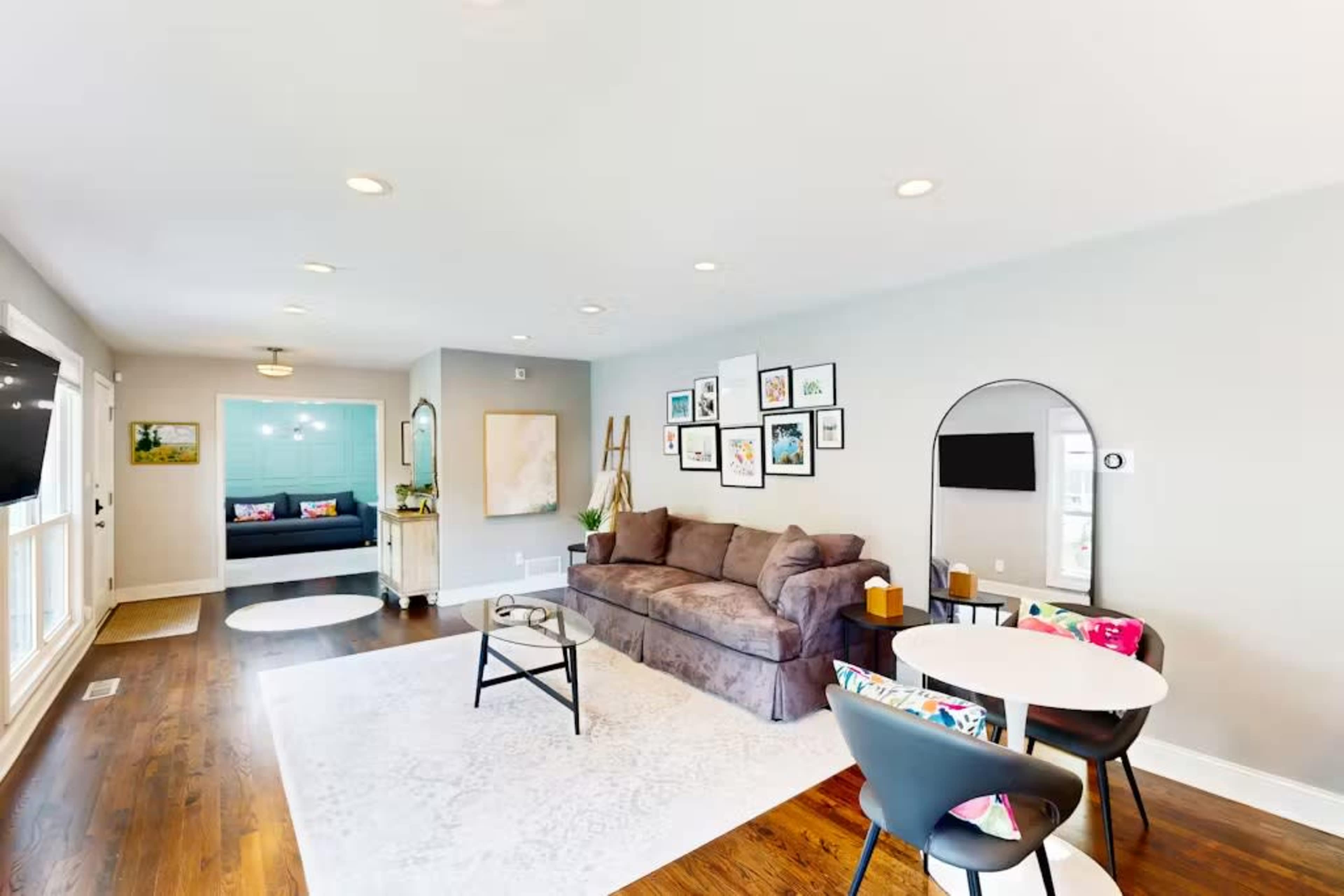 The image shows a modern living room with a brown sectional sofa, a circular dining table with black chairs, and various framed artworks on the walls.