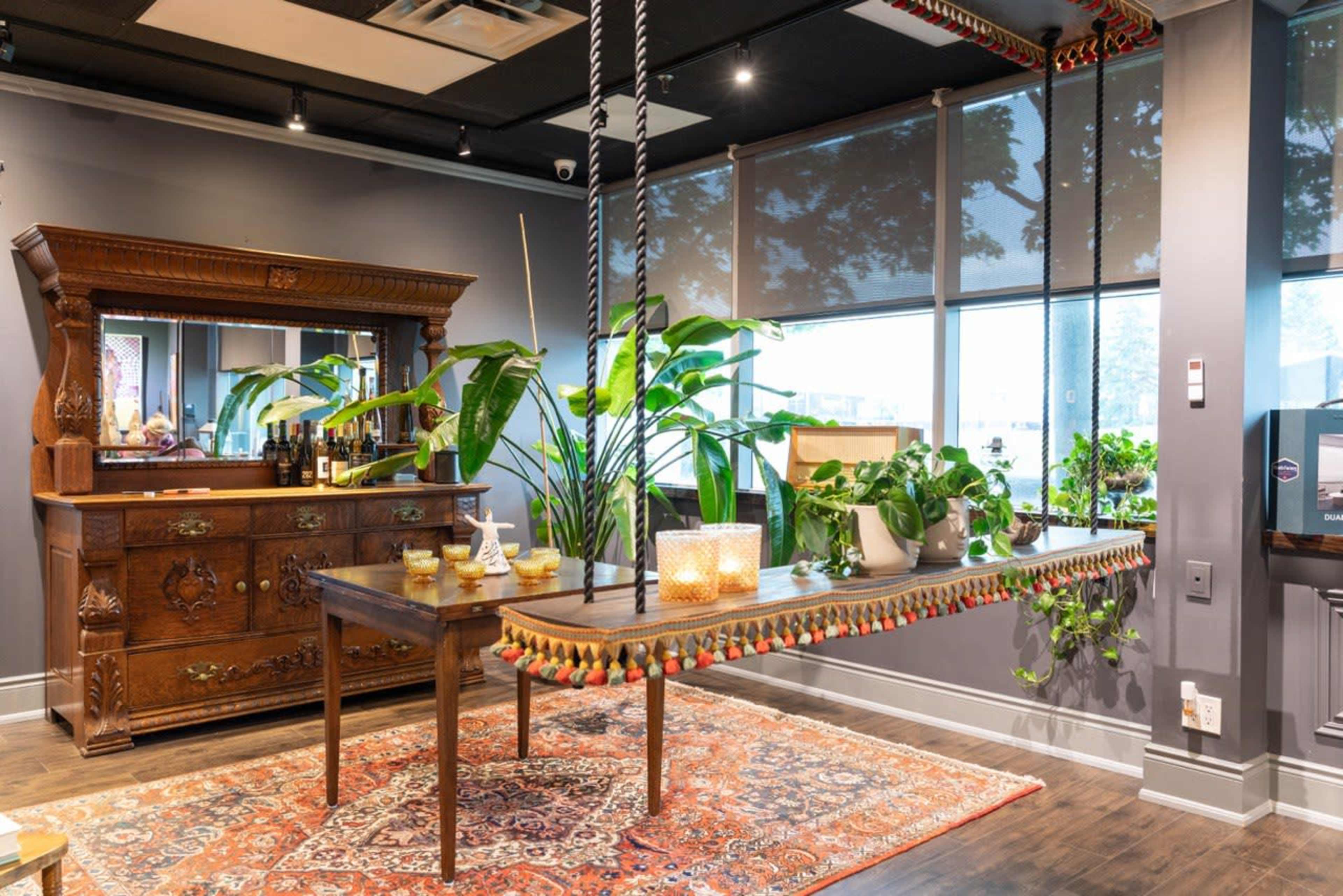 The image shows a modern interior space featuring a wooden table with decorative trim, an ornate dresser, large indoor plants, and large windows allowing natural light.