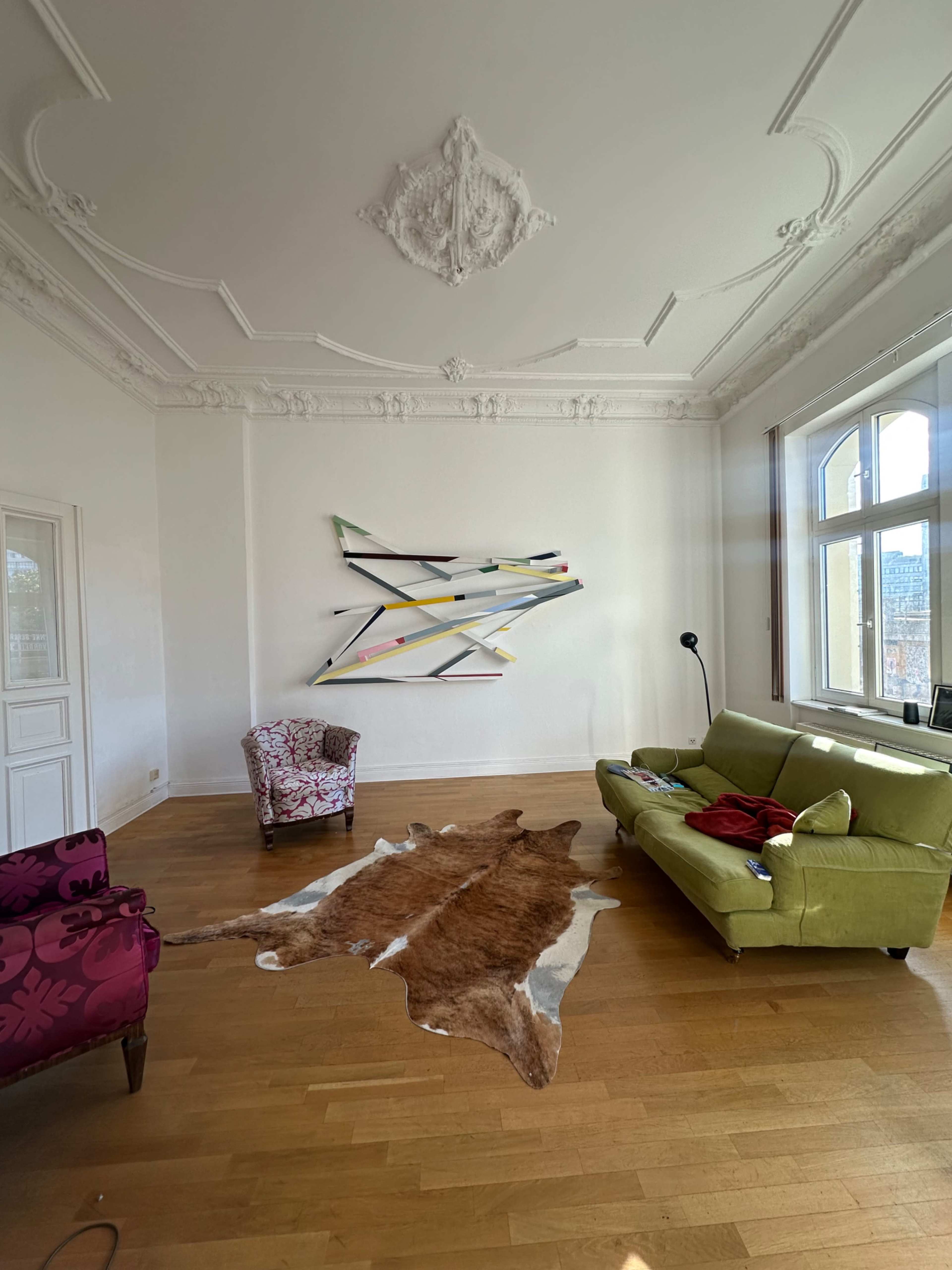 The image shows a spacious living room featuring a green sofa, a patterned armchair, a cowhide rug on the wooden floor, and abstract wall art above.