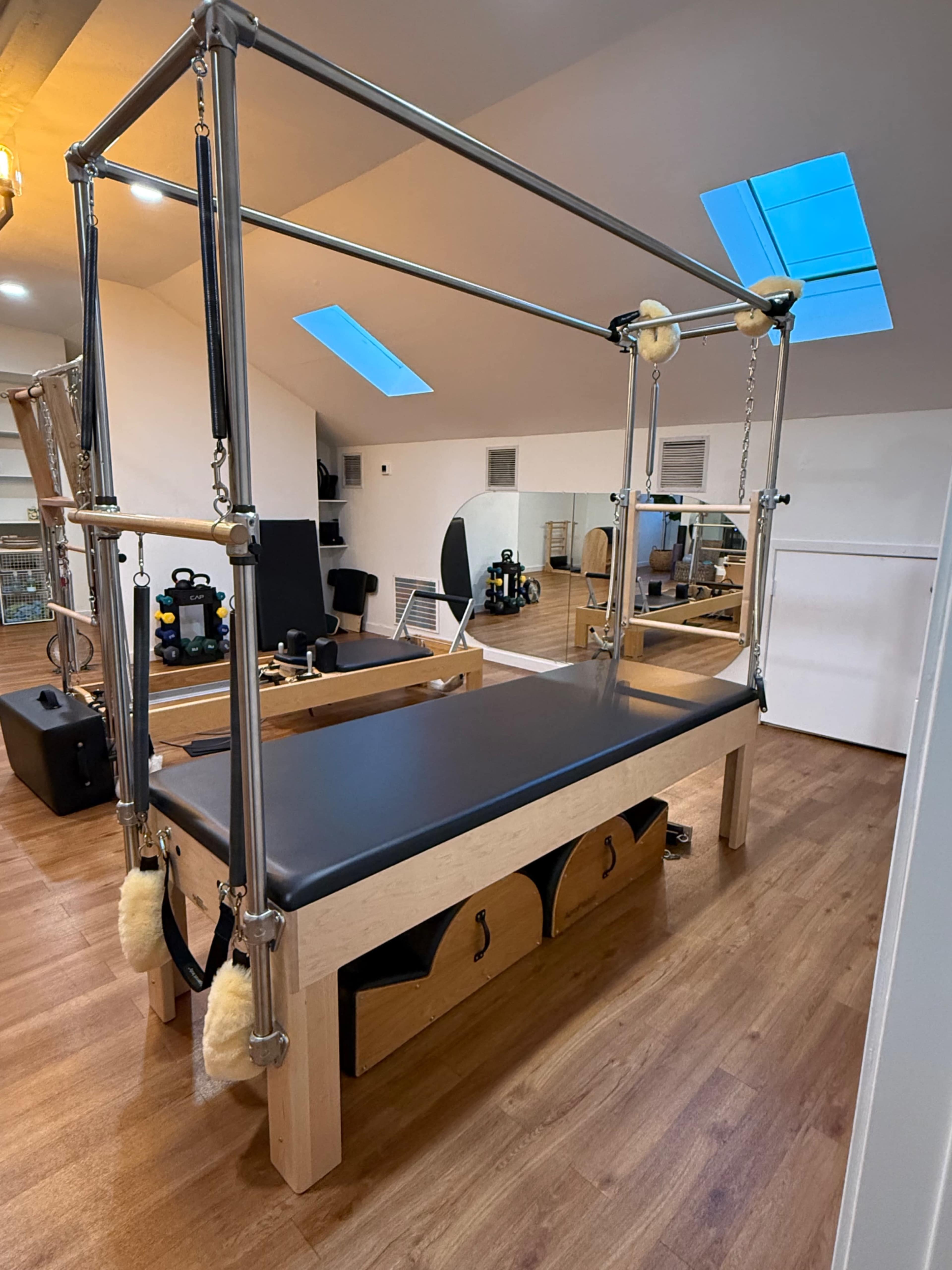 The image shows a Pilates reformer equipped with a wooden base and metal framework, situated in a well-lit exercise studio with wooden flooring and various fitness equipment in the background.