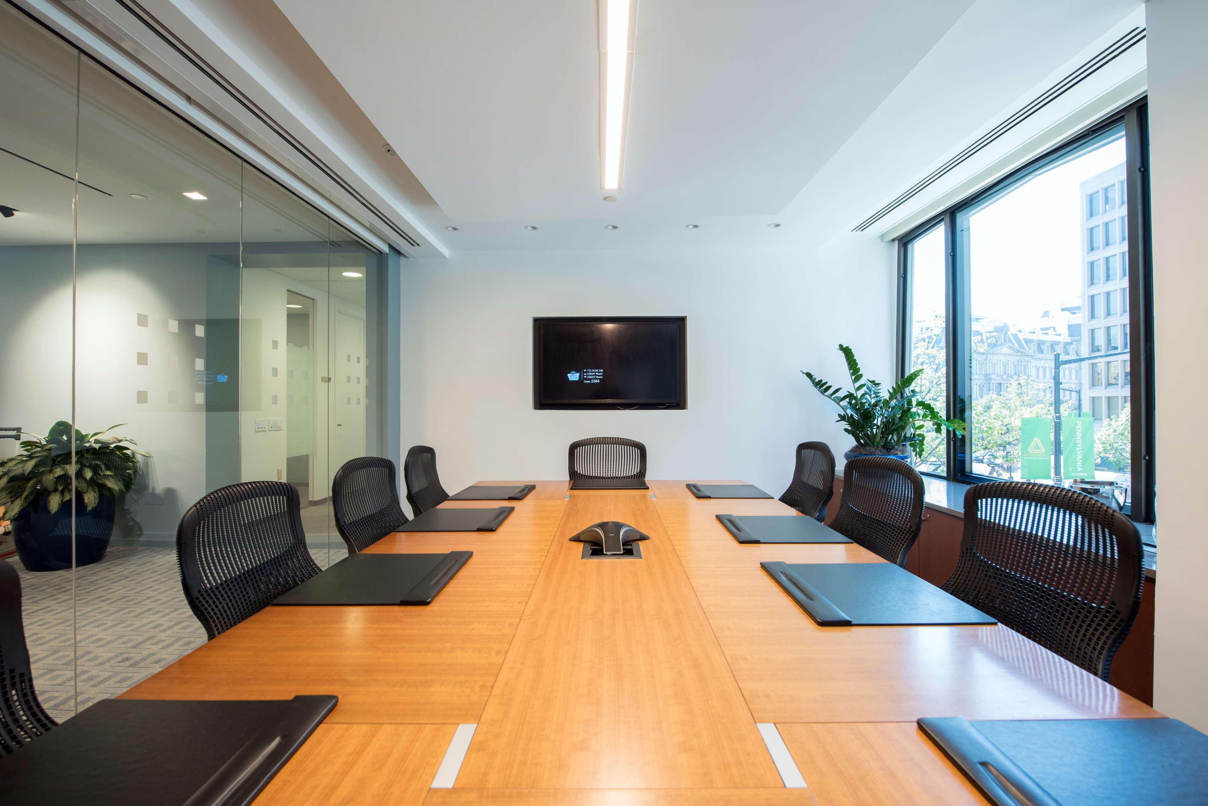 A modern conference room features a large wooden table surrounded by black chairs, with a television mounted on the wall and windows offering a view of the outside.