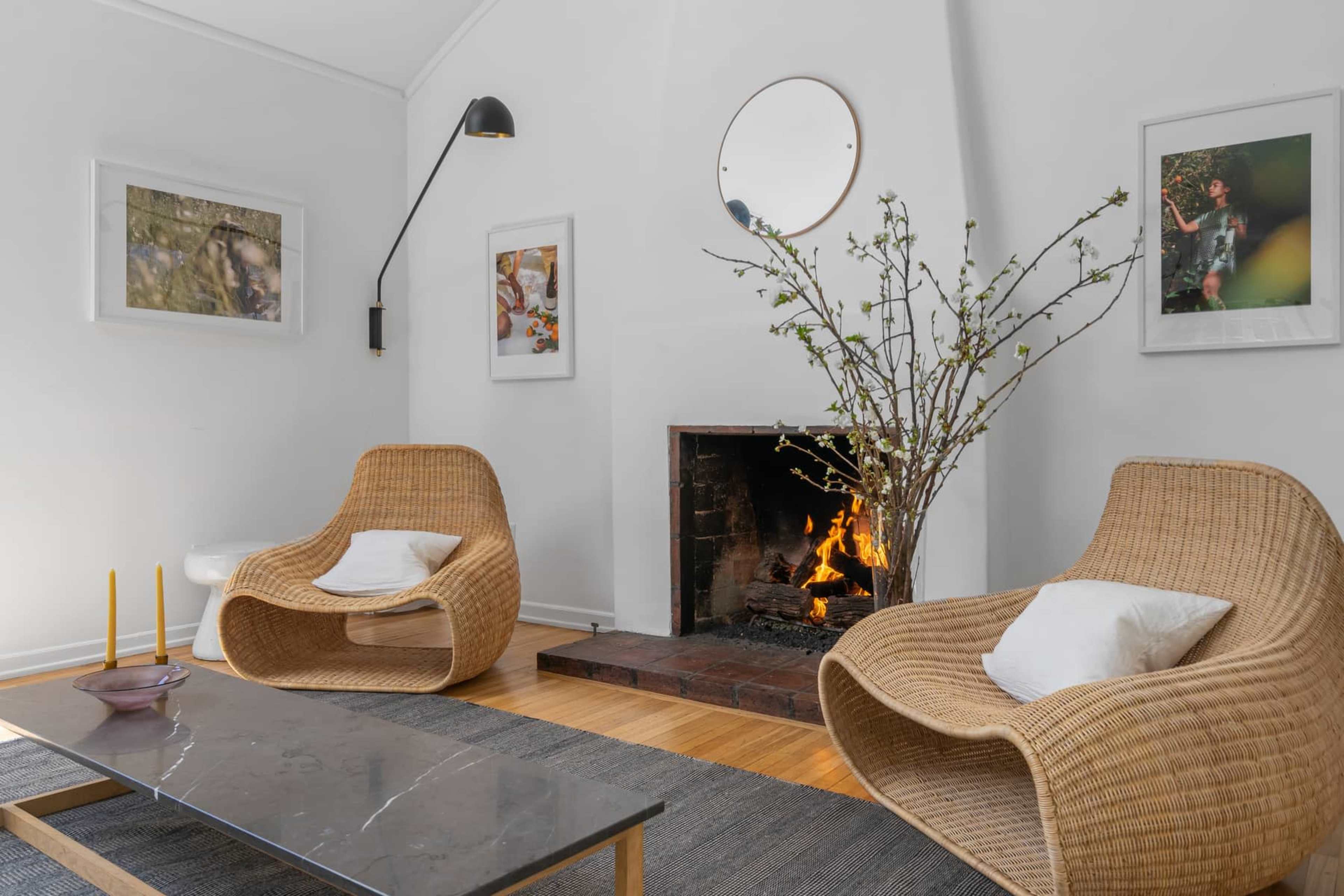 The scene features two woven chairs positioned near a fireplace, which contains a lit fire, alongside a coffee table and decorative wall art.