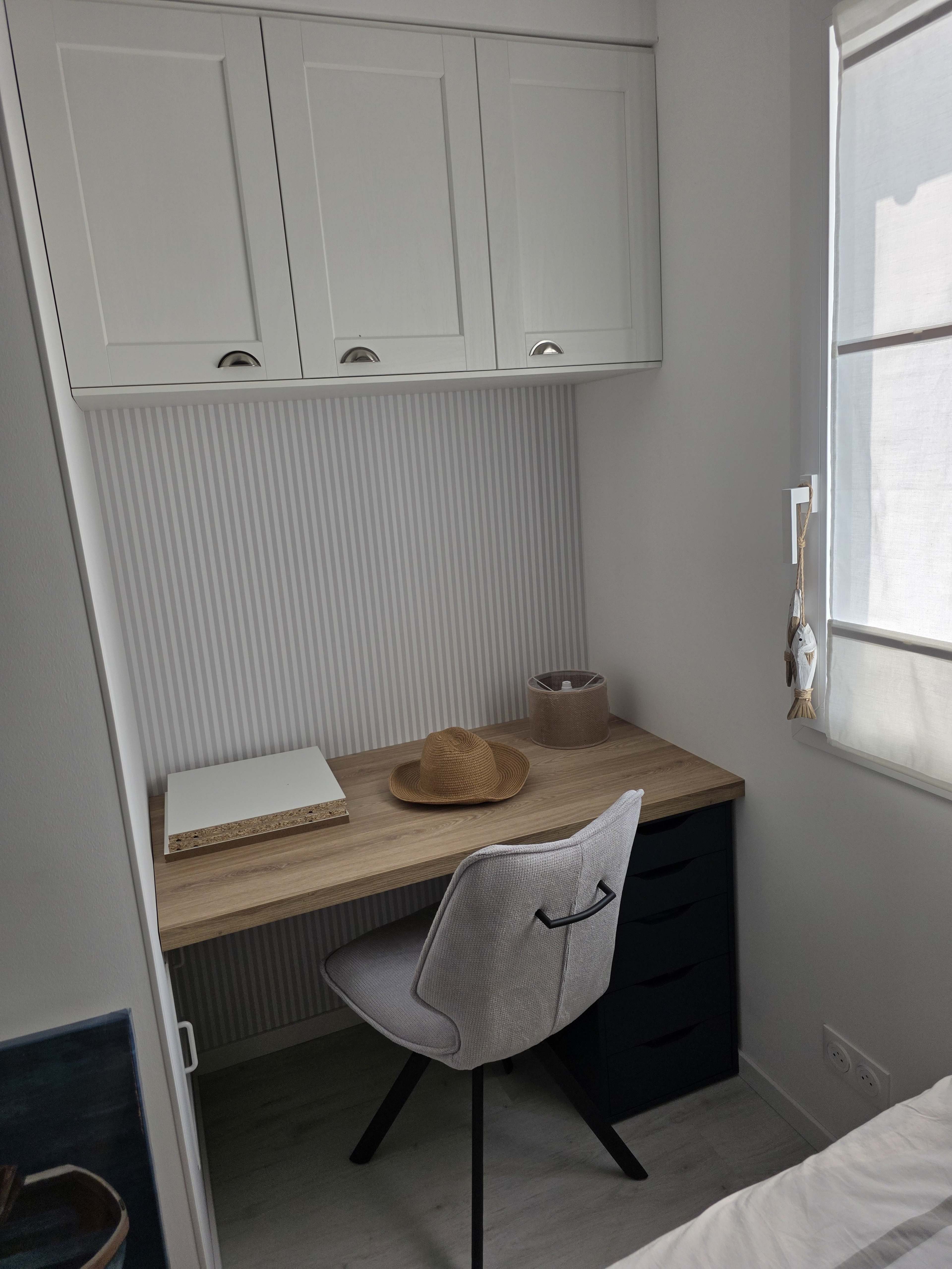 The image shows a small, minimalist home office setup featuring a wooden desk with a chair, a hat, and a few decorative items against a white wall with cabinets above.