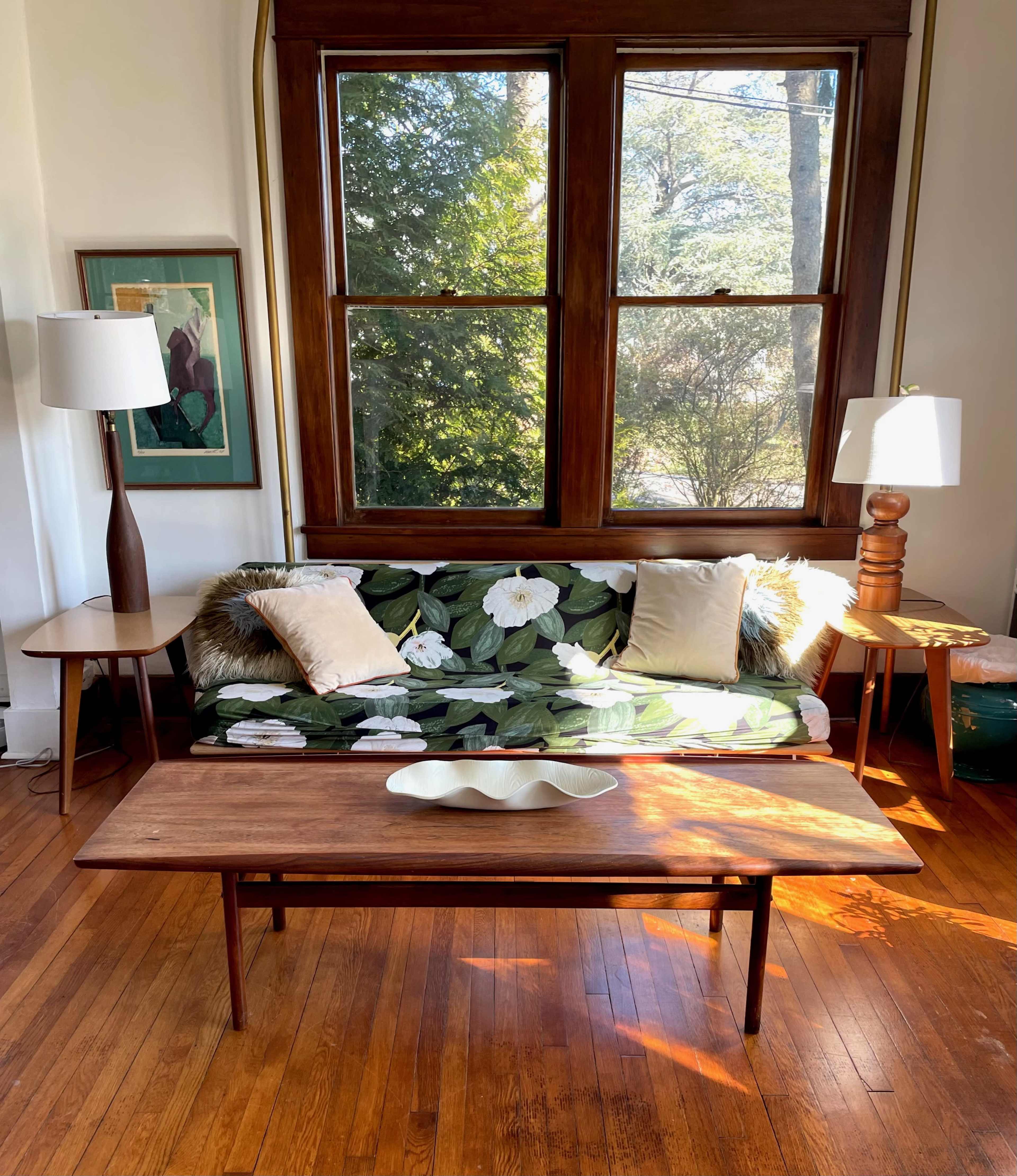The scene features a living room with a floral-patterned sofa, two lamps, and a wooden coffee table under large windows letting in natural light.
