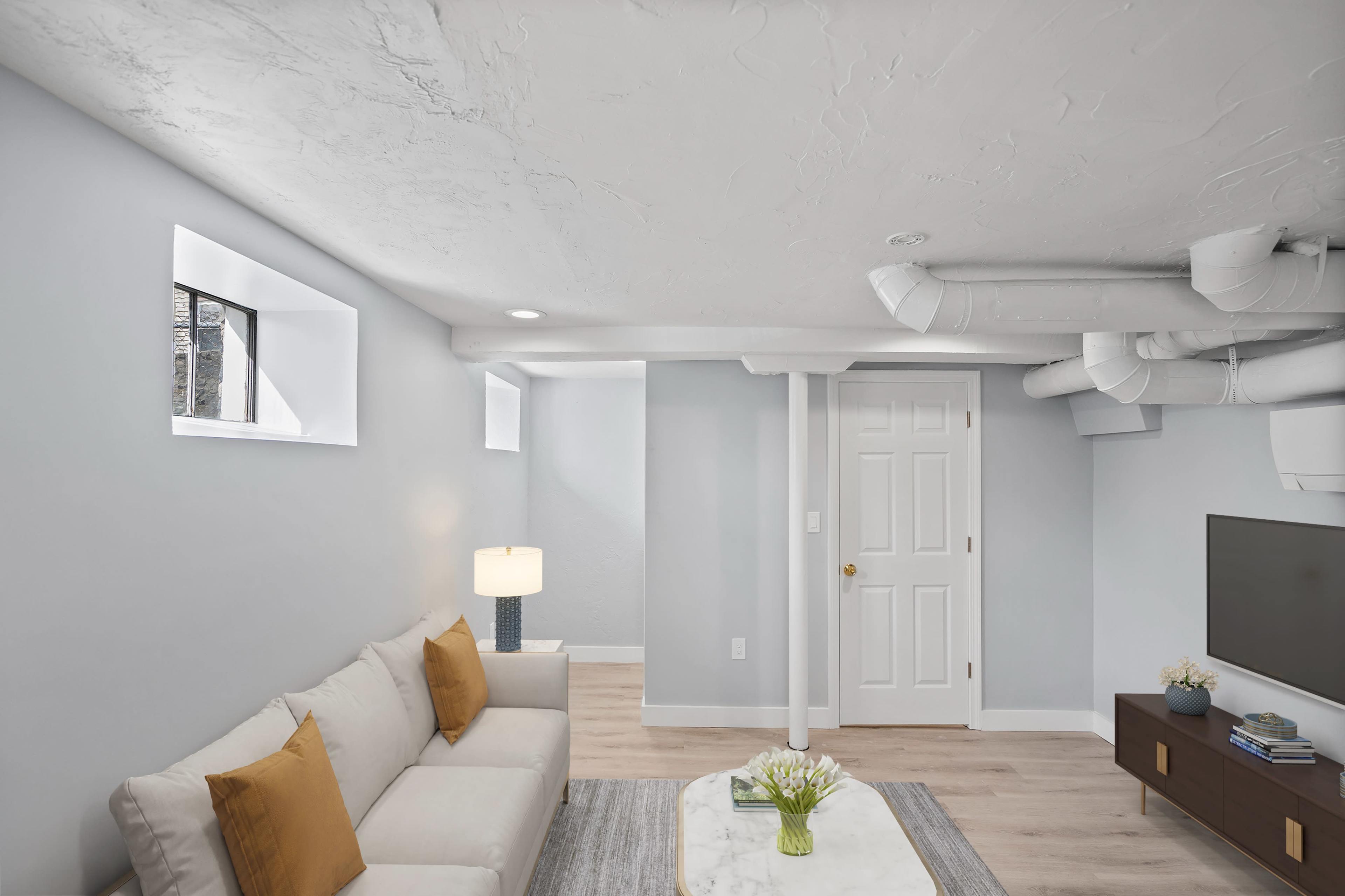A modern basement living area with a light-colored couch, a small table with flowers, a television on a stand, and a door in the background.