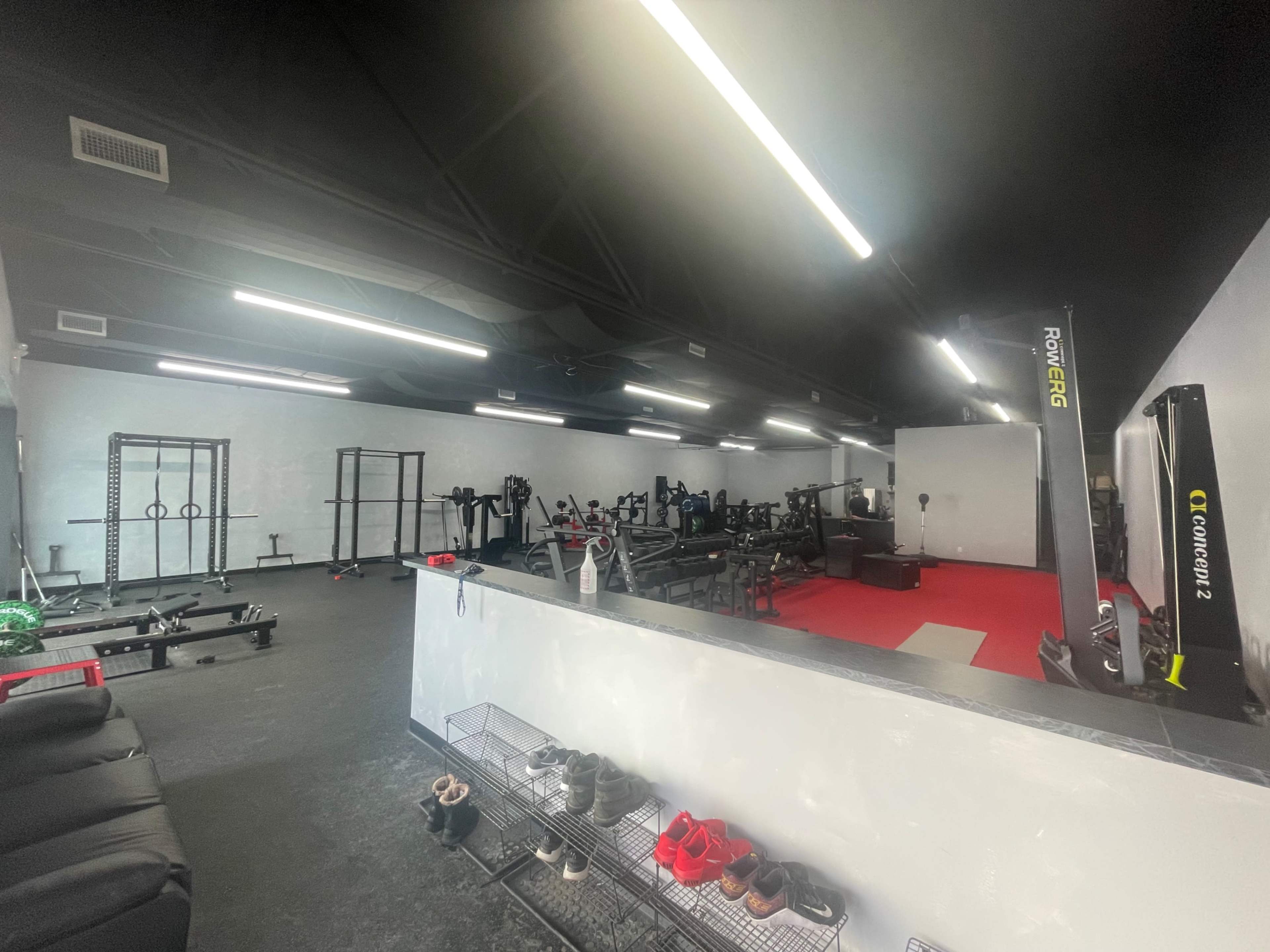 A fitness gym with various exercise equipment, including weight racks and rowing machines, set against a backdrop of dark walls and bright overhead lights.