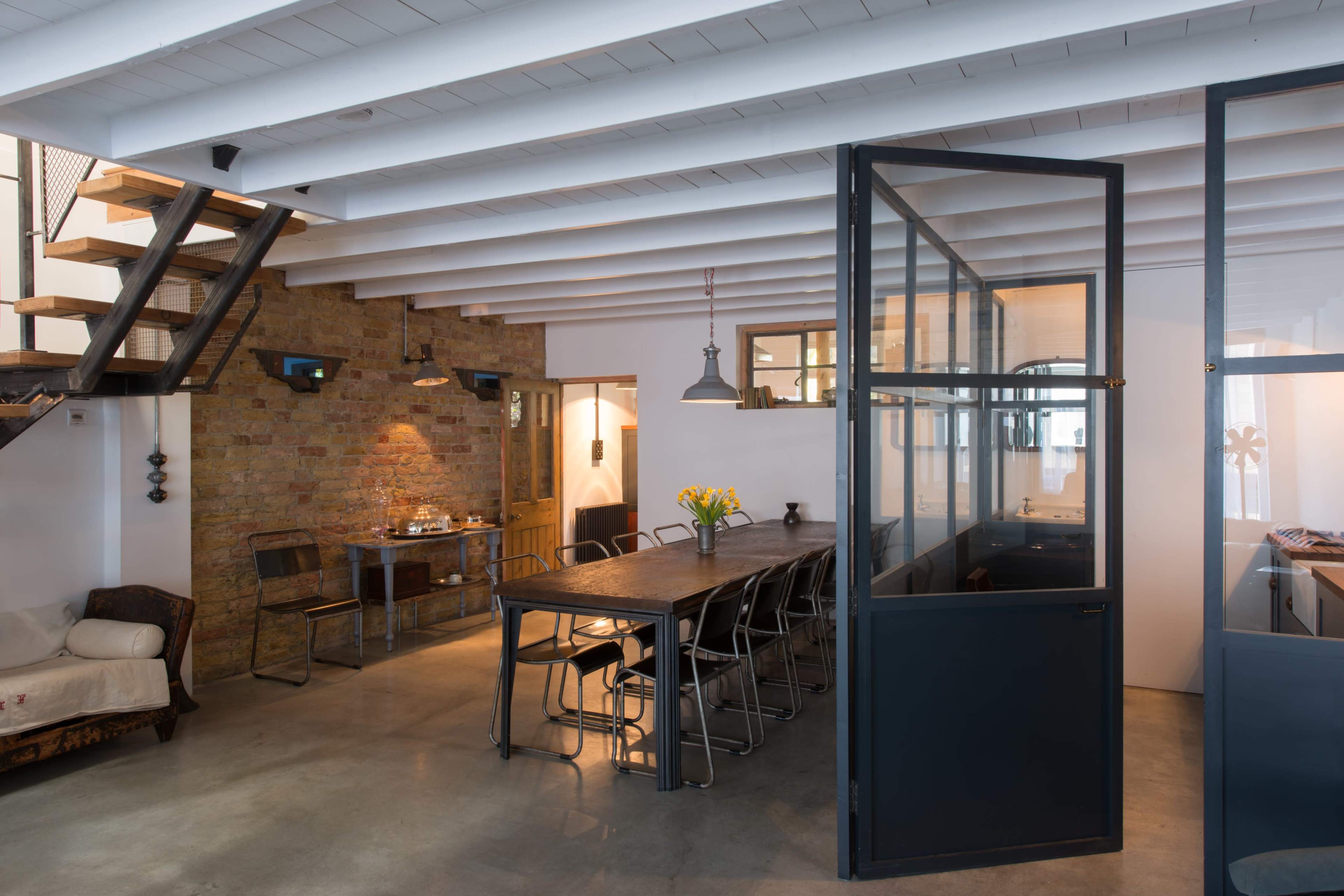 The image shows a modern, open-concept space featuring a dining area with a long table and chairs, a staircase to the left, and glass partitions separating different sections.