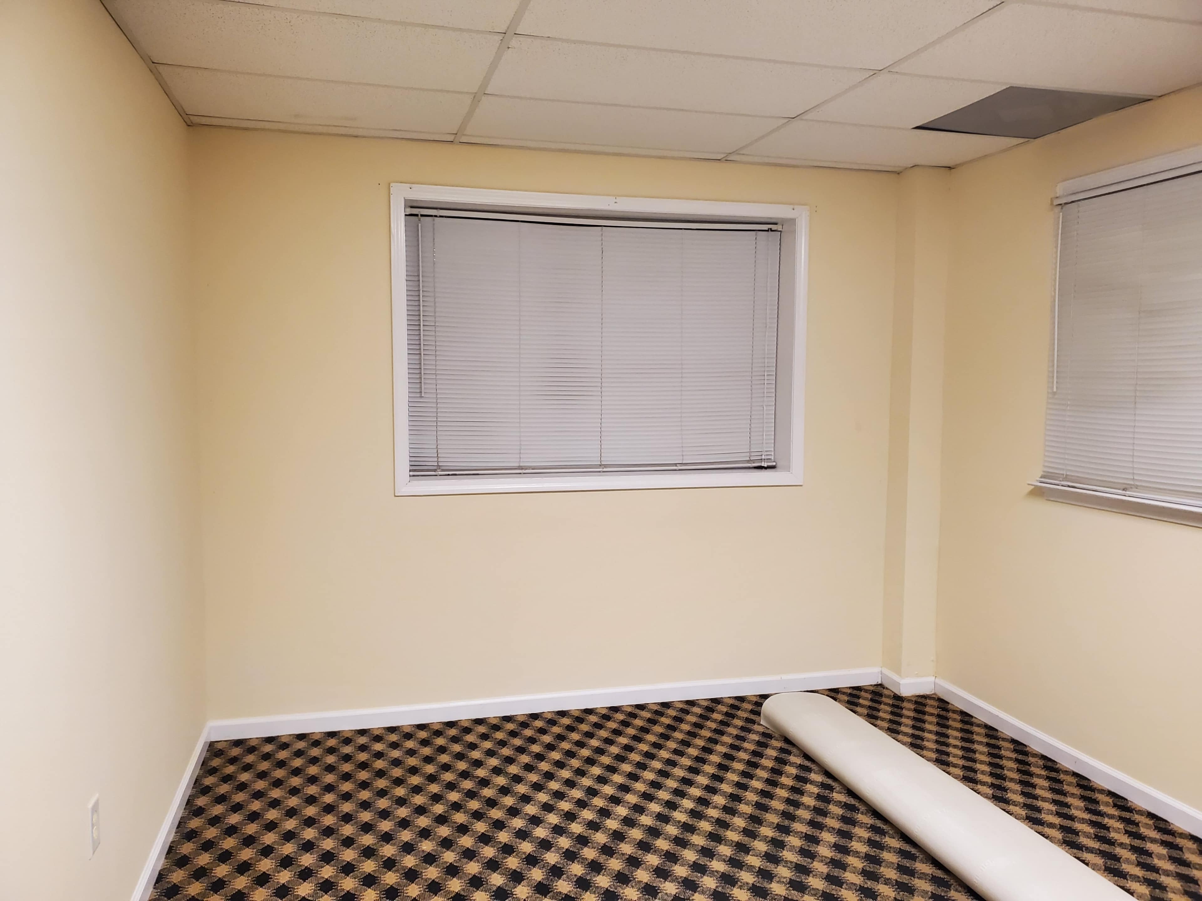 A small, empty room with beige walls, a window covered by blinds, and a rolled-up carpet on the floor.