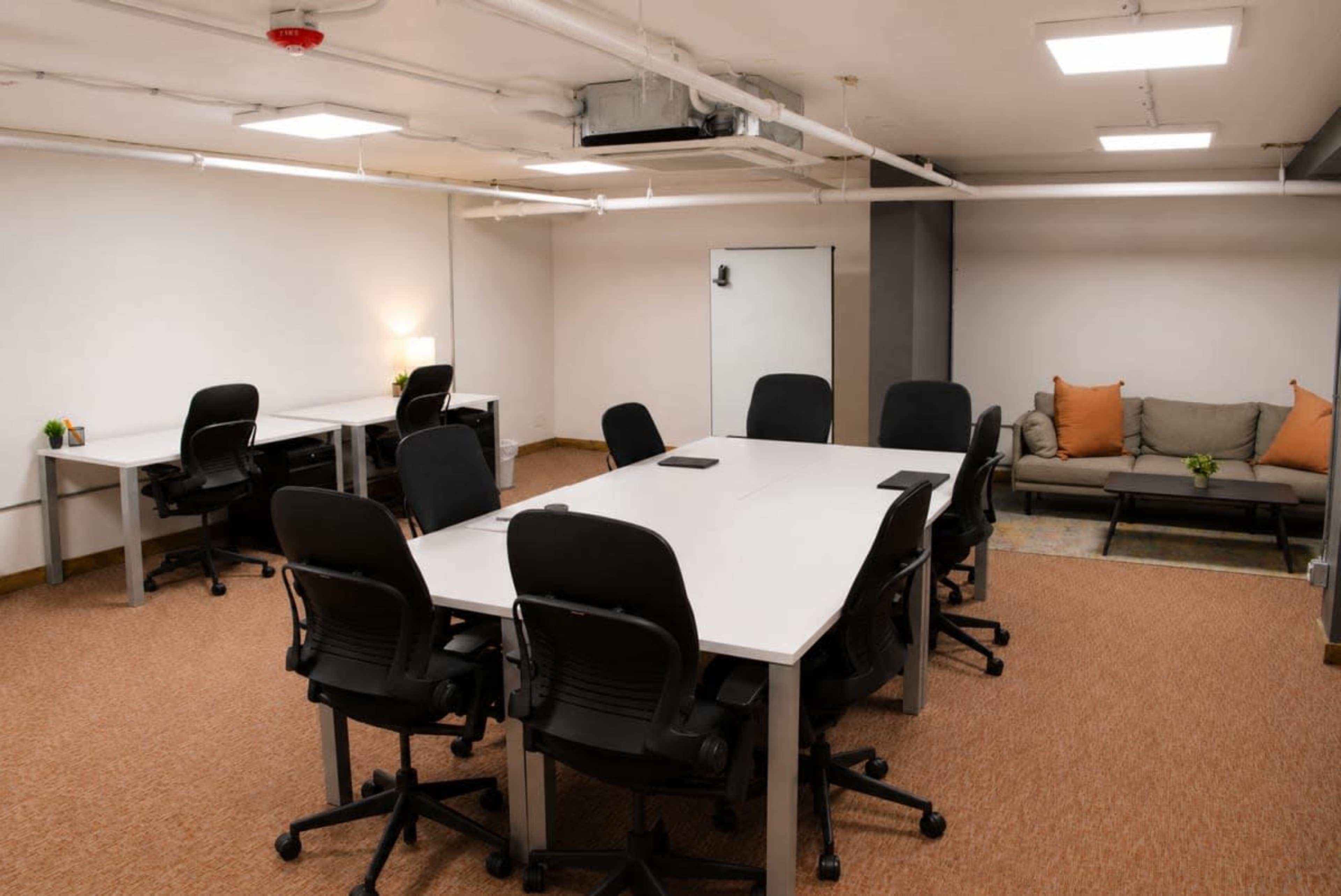 The image depicts a modern conference room with a large table surrounded by chairs, additional desks, and a seating area with a couch and cushions.