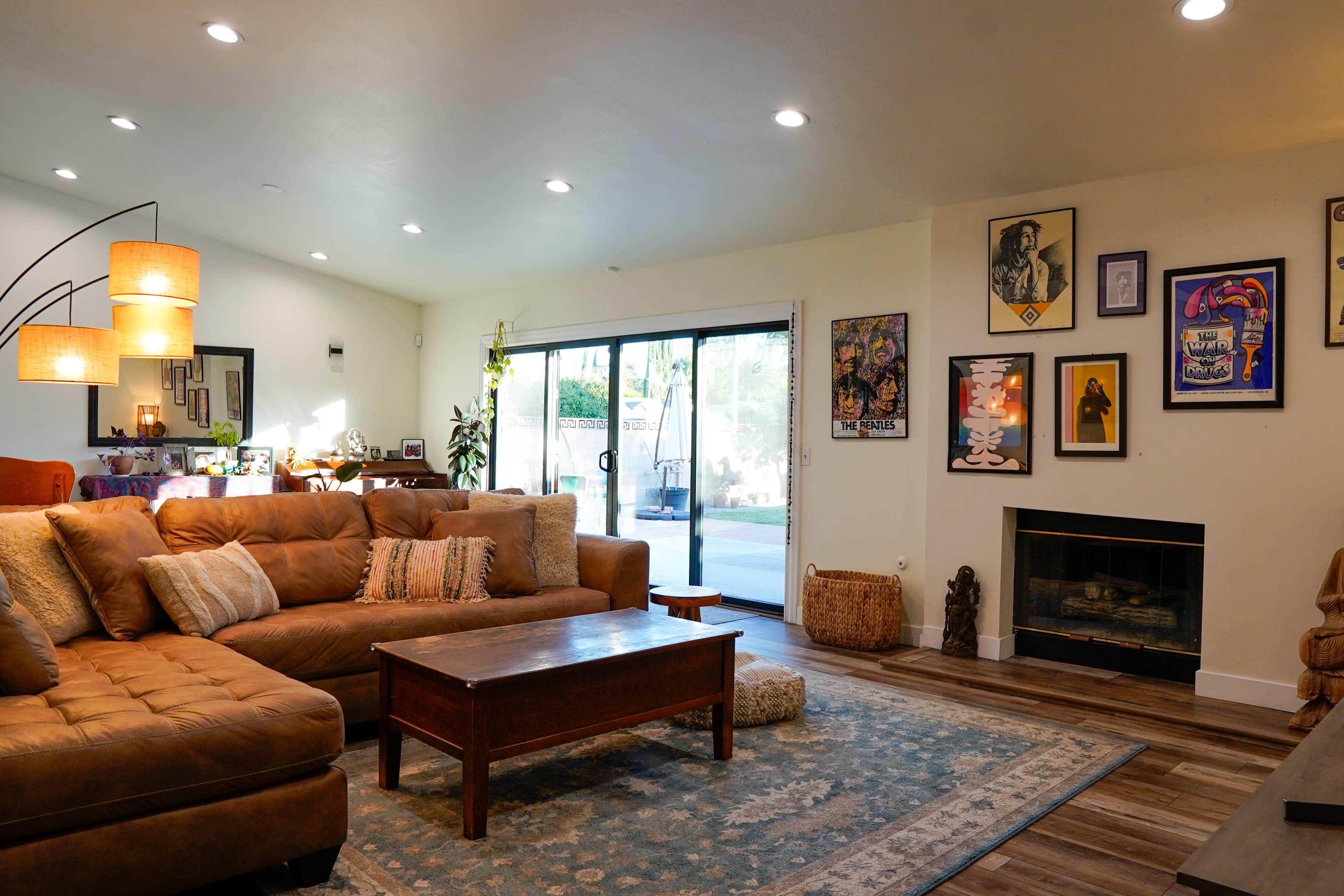 The living room features a large brown sectional couch, a wooden coffee table, and several framed artworks on the walls, with a glass door leading to a patio area.