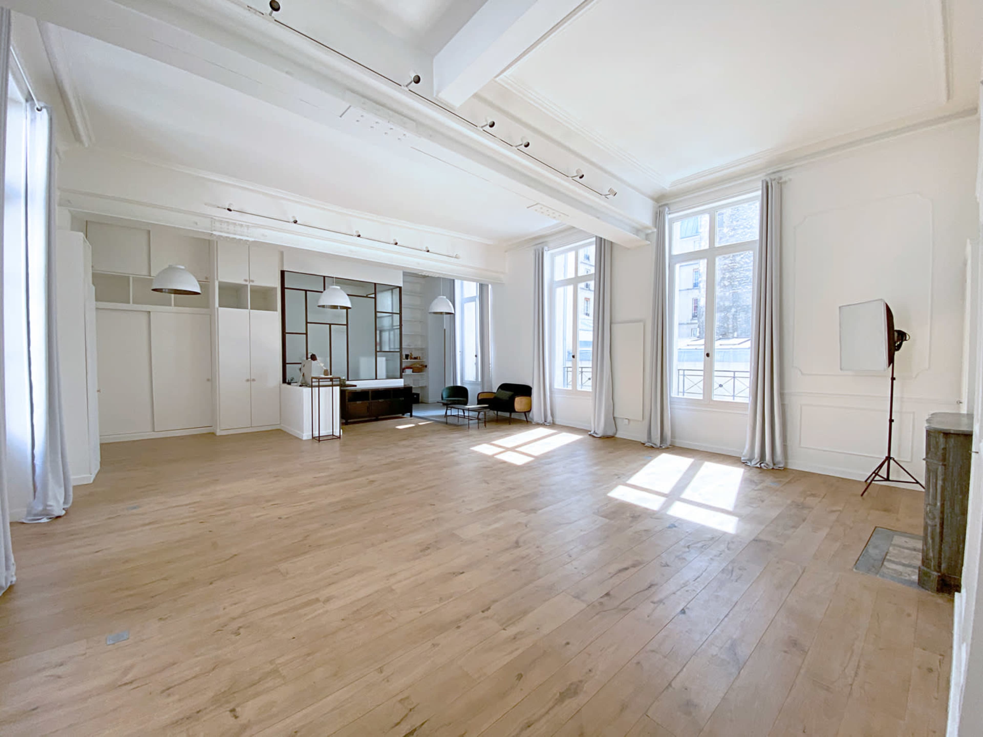 The image shows a spacious, well-lit room with wooden flooring, large windows, and minimal furnishings.