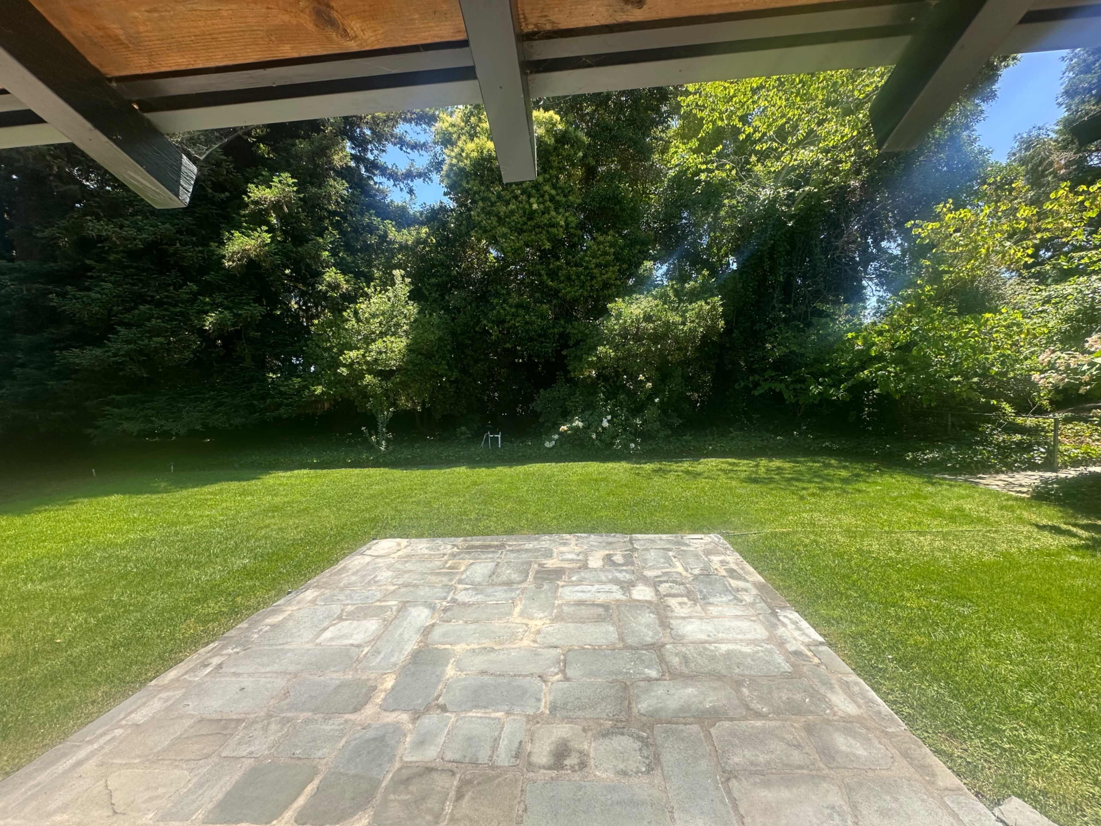 A stone patio is visible in the foreground, leading to a lush green lawn surrounded by trees and shrubs.