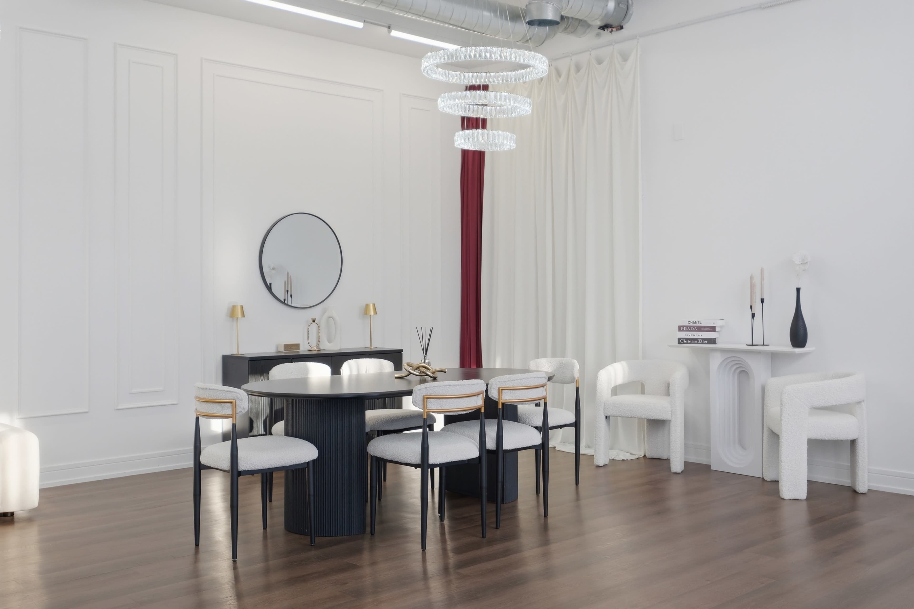 The image shows a modern interior space featuring a round black dining table surrounded by six chairs, a circular mirror on the wall, and a decorative light fixture overhead, with white curtains and minimalist decor elements.