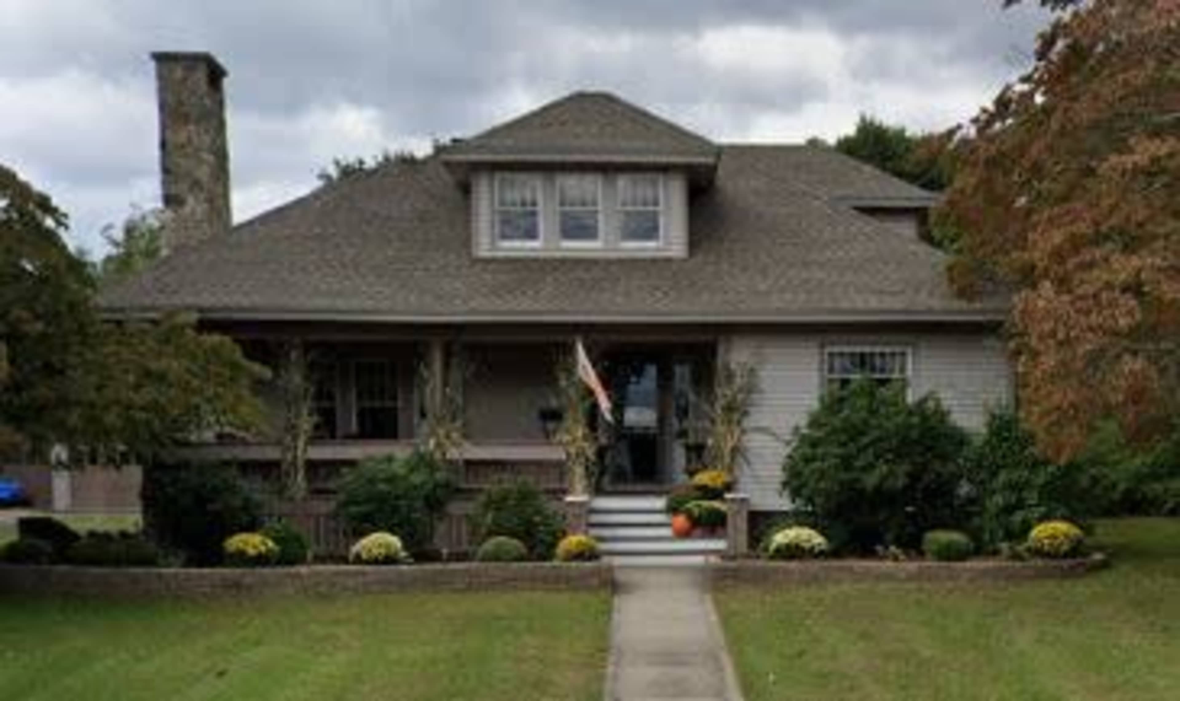 A large, two-story house with a front porch, flanked by trees and decorative plants.