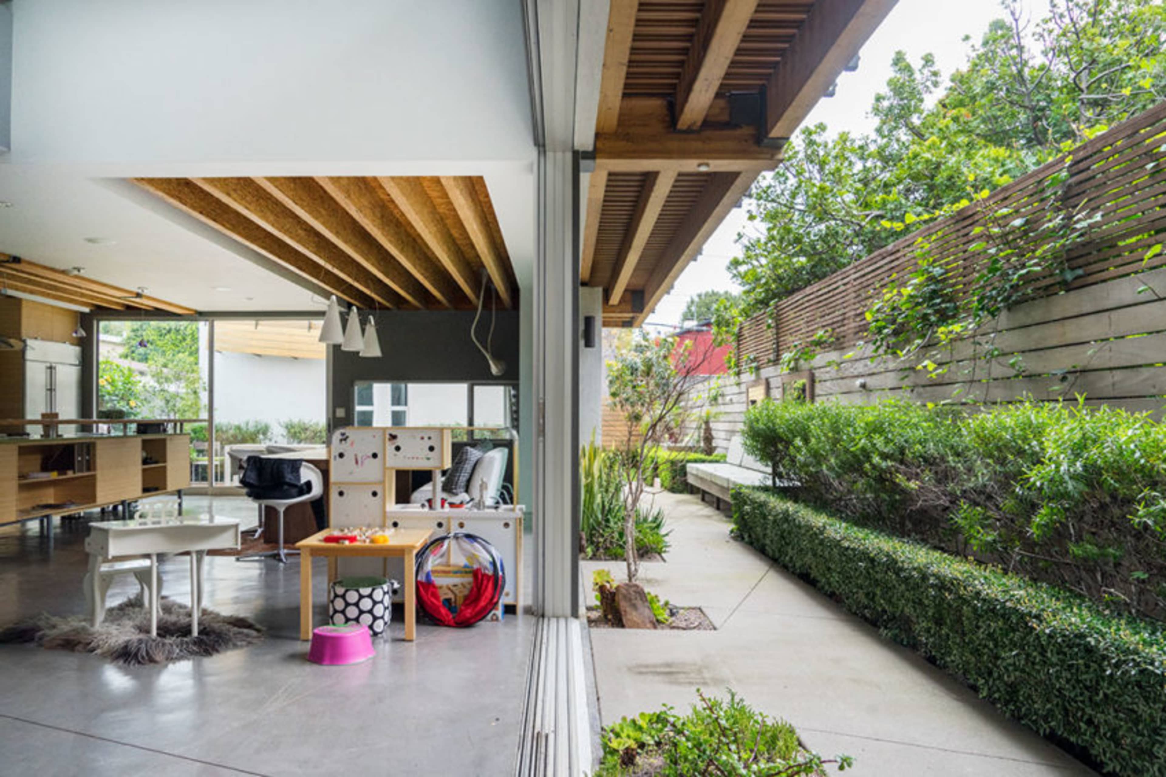 The image shows a modern interior space with a dining area and play area adjacent to an outdoor garden with neatly trimmed hedges.