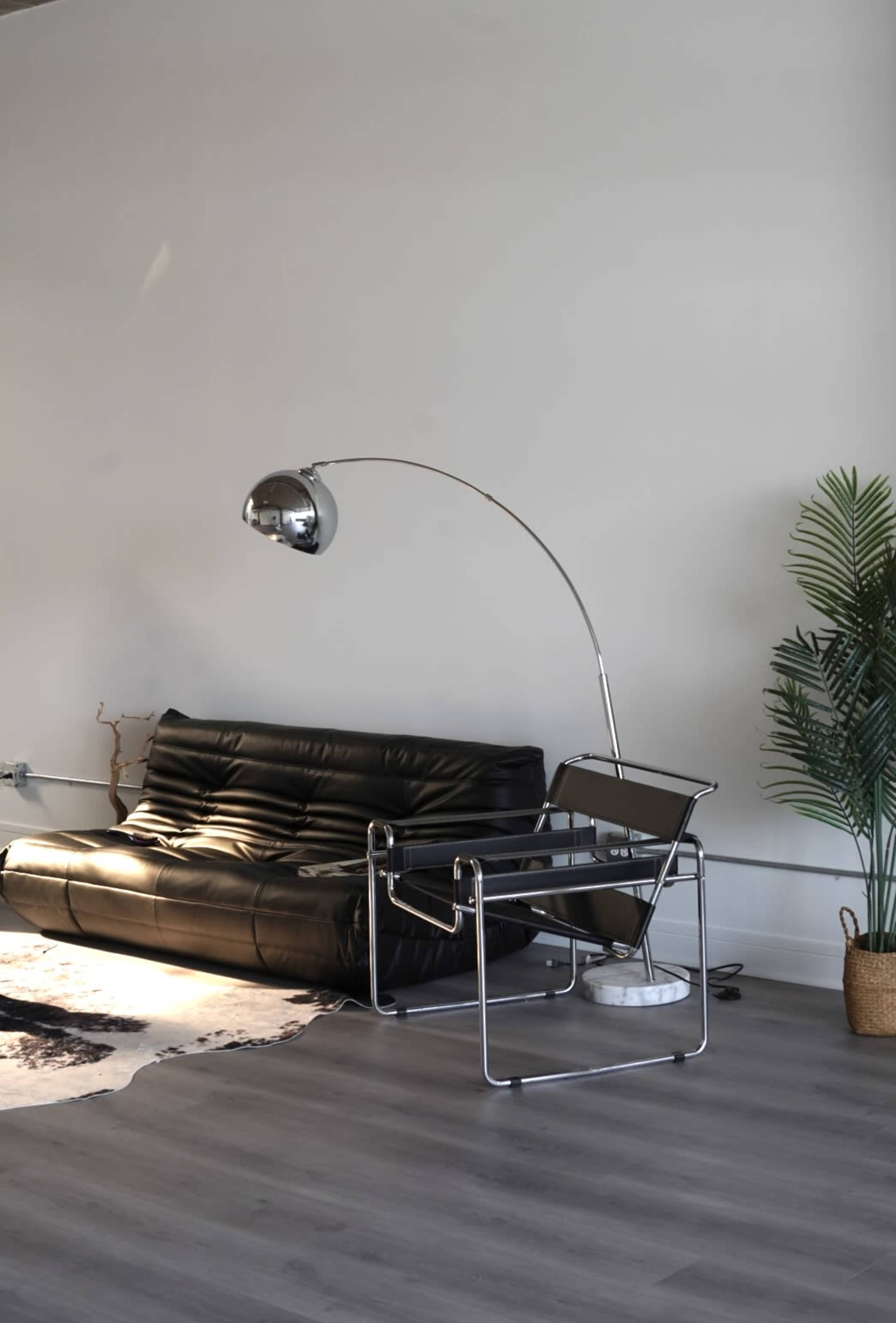 The scene features a modern living space with a black leather sofa, a sleek arc floor lamp, a contemporary chair, and a potted plant.