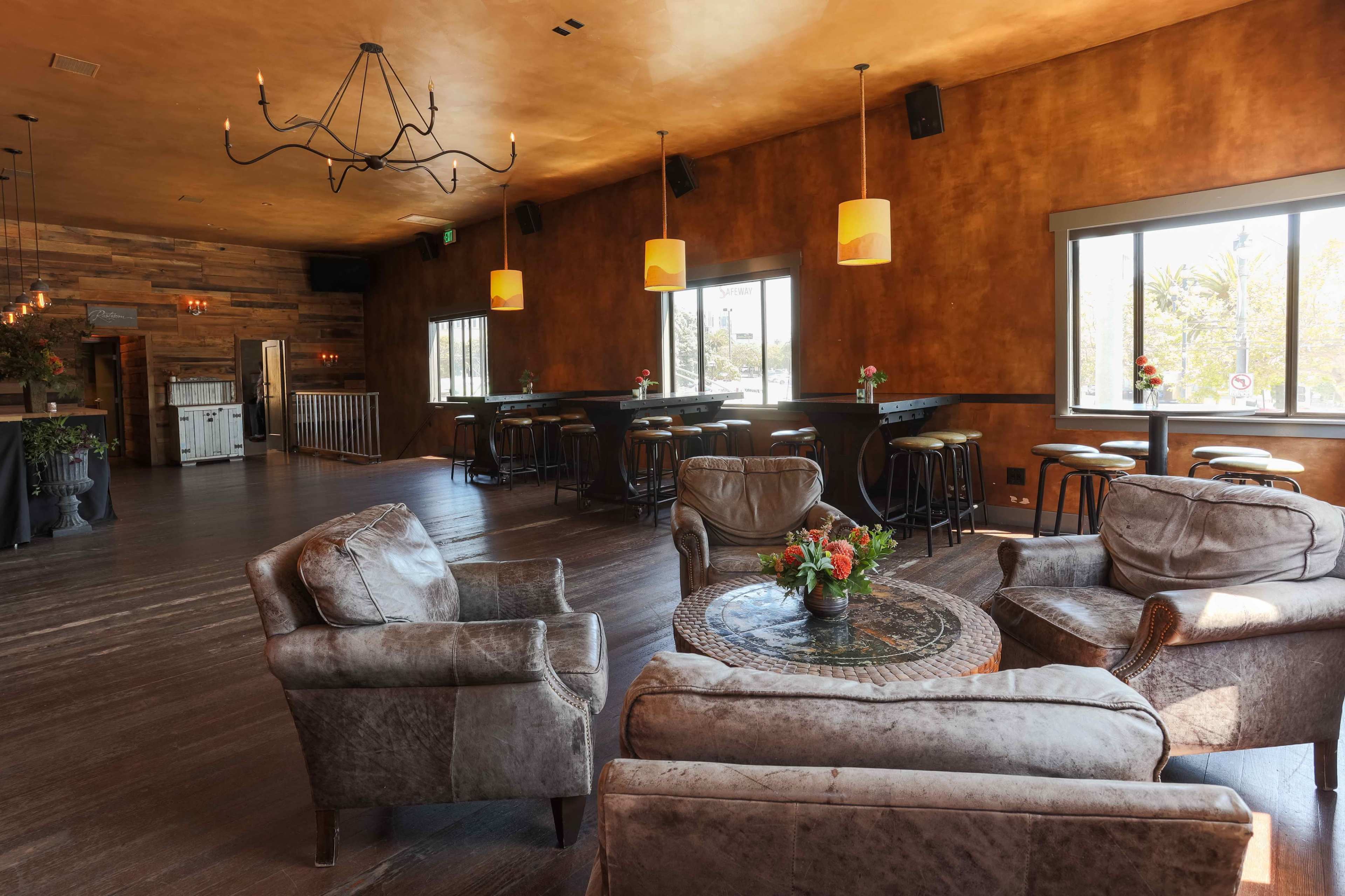 A restaurant interior with a seating area featuring plush armchairs around a circular table, large windows, and a bar area with high stools.