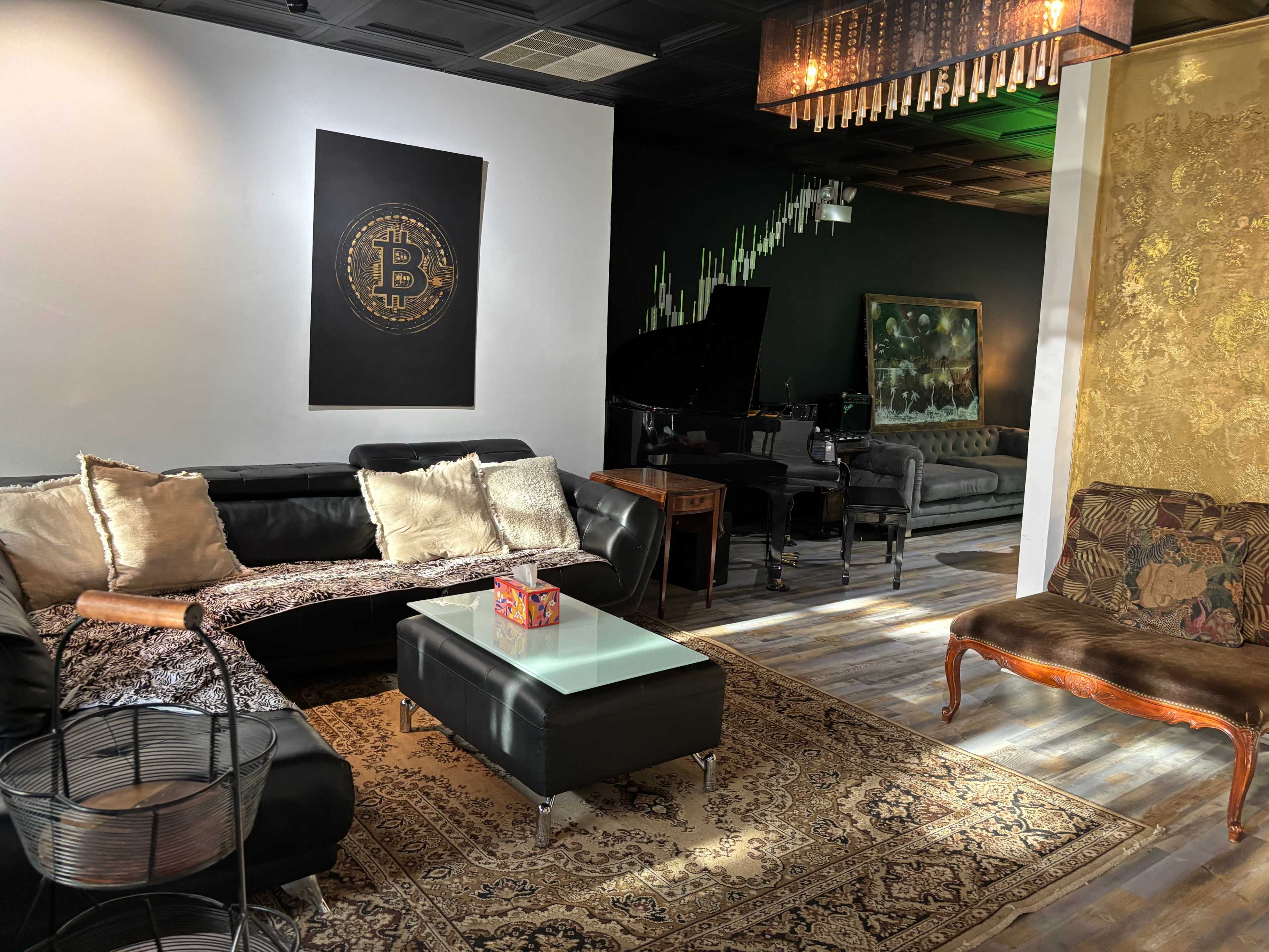 The image shows a stylish lounge area with black sofas, a glass coffee table, and decorative elements like a chandelier, alongside a grand piano and various artworks on the walls.