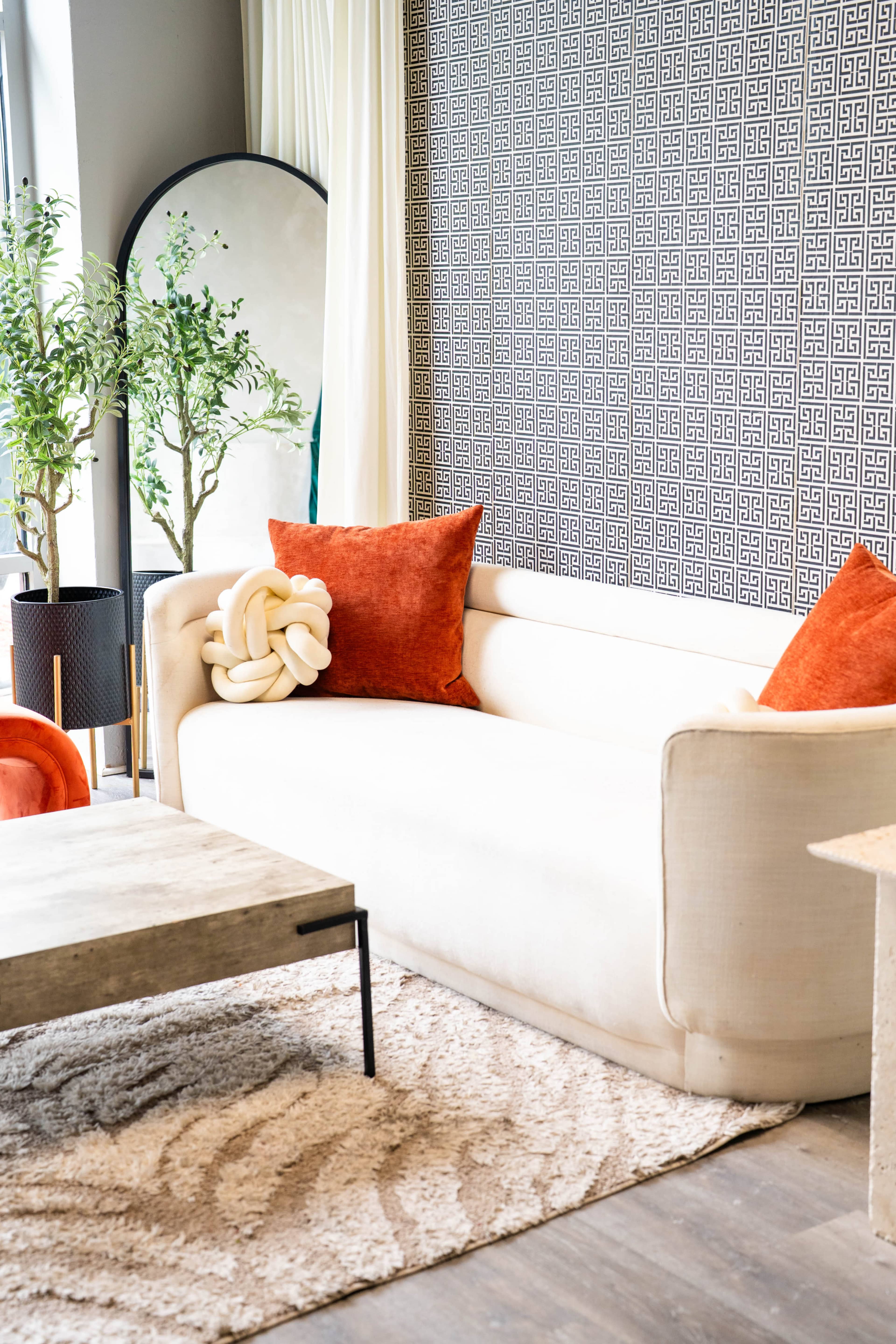 The image shows a modern living room with a beige sofa, orange accent pillows, a round mirror, and a wooden coffee table on a textured rug.