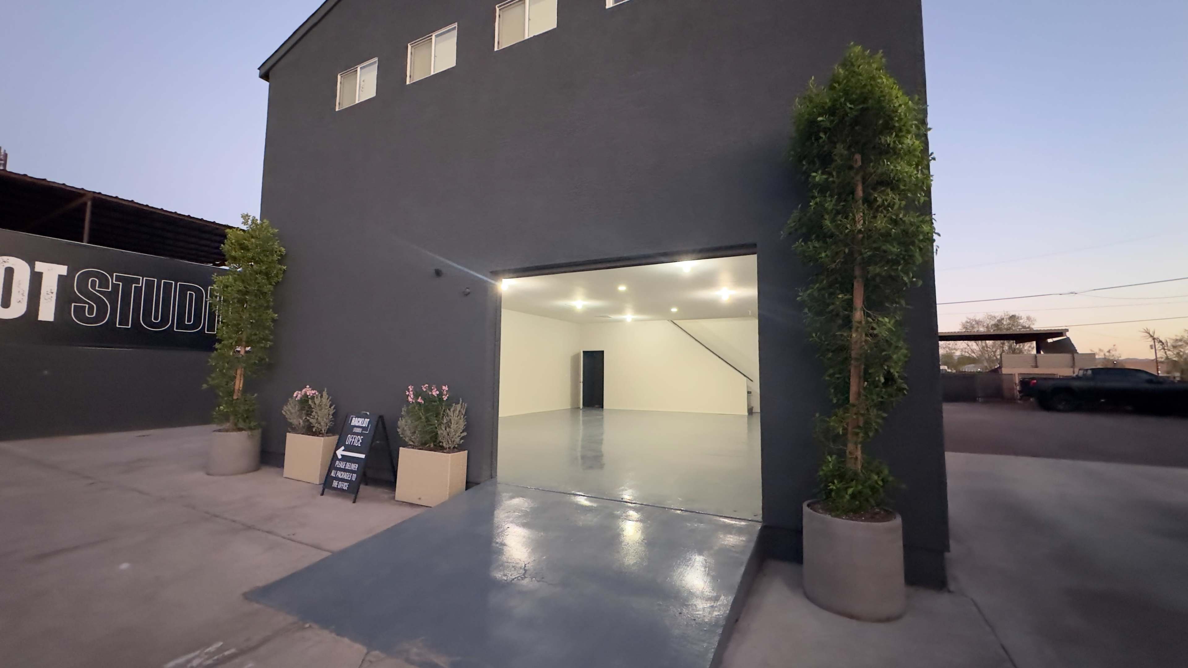 A modern, two-story building with a large open garage entrance, flanked by planters and illuminated by overhead lights.