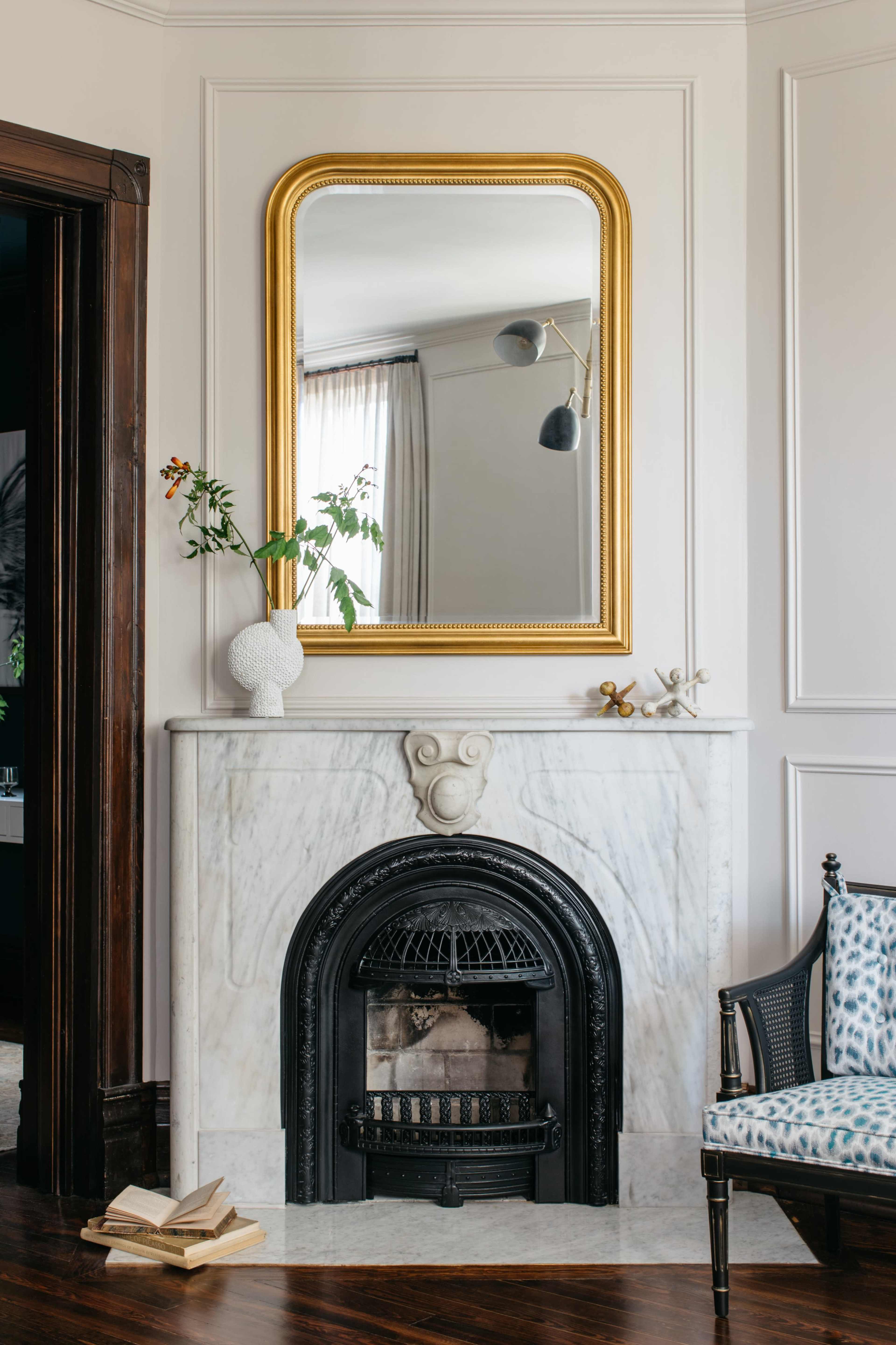 A marble fireplace with a mirrored frame and decorative elements is set against a wall in a room featuring a chair and books on the floor.