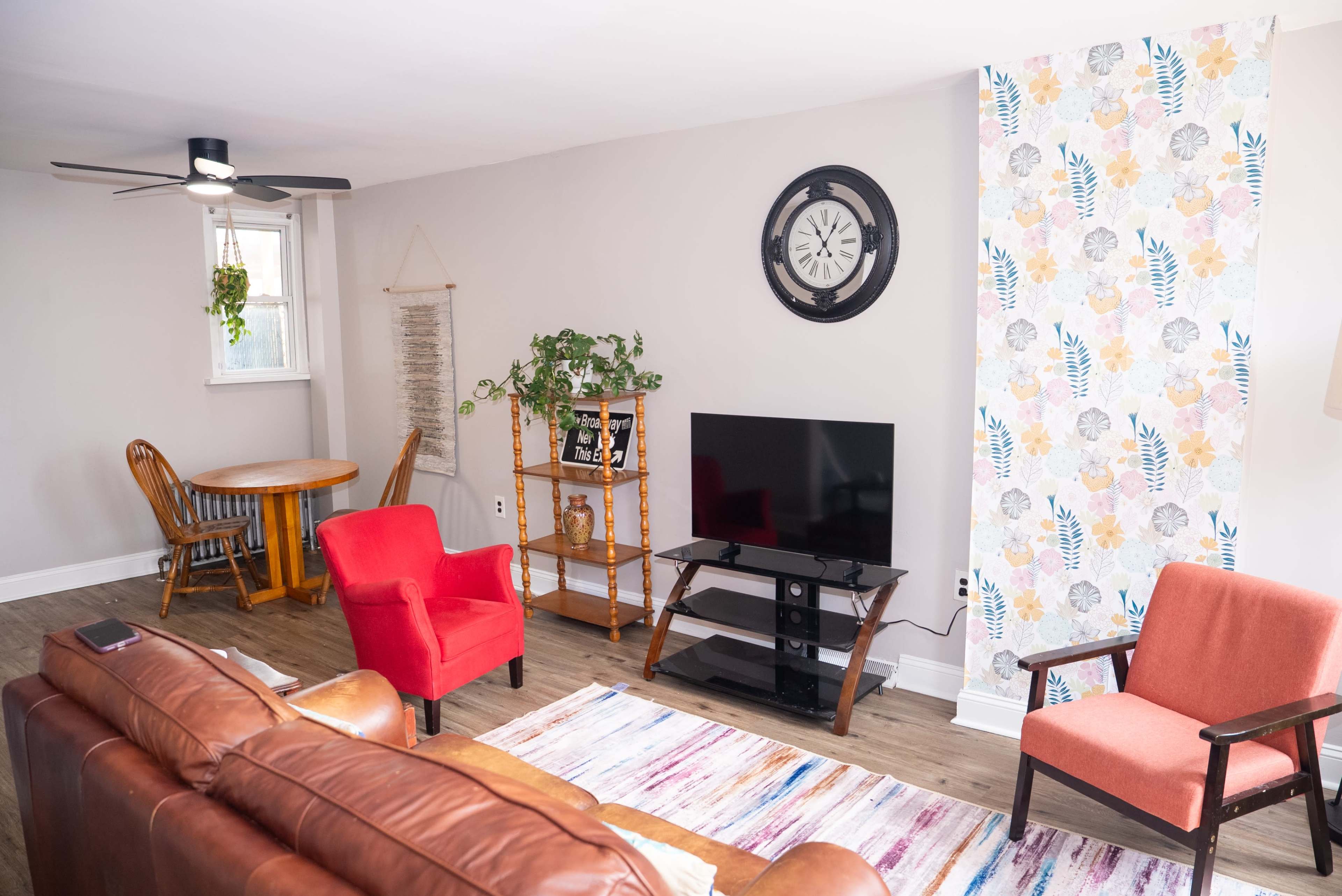 The image shows a living room featuring a brown leather sofa, a red armchair, a wooden dining table, and a television on a black stand, with a floral accent wall and a ceiling fan.