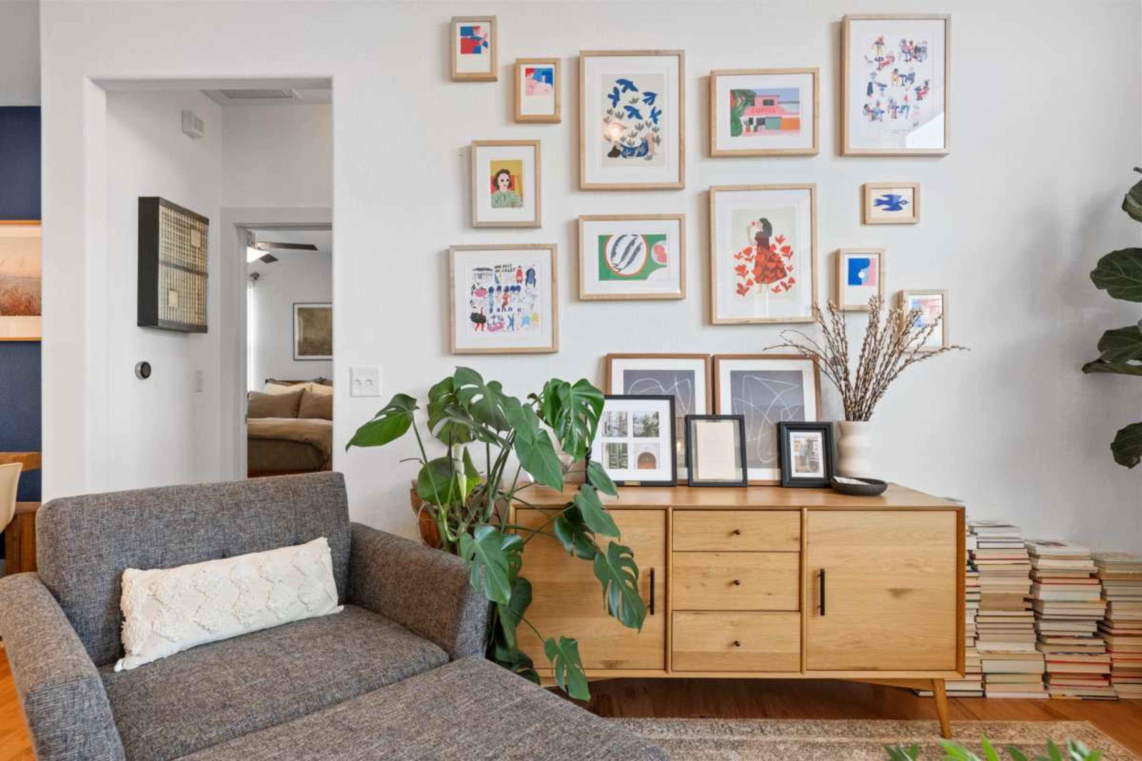 The image features a living room corner with a gray couch, a wooden sideboard adorned with framed artwork, and a houseplant beside a gallery wall of framed pictures.