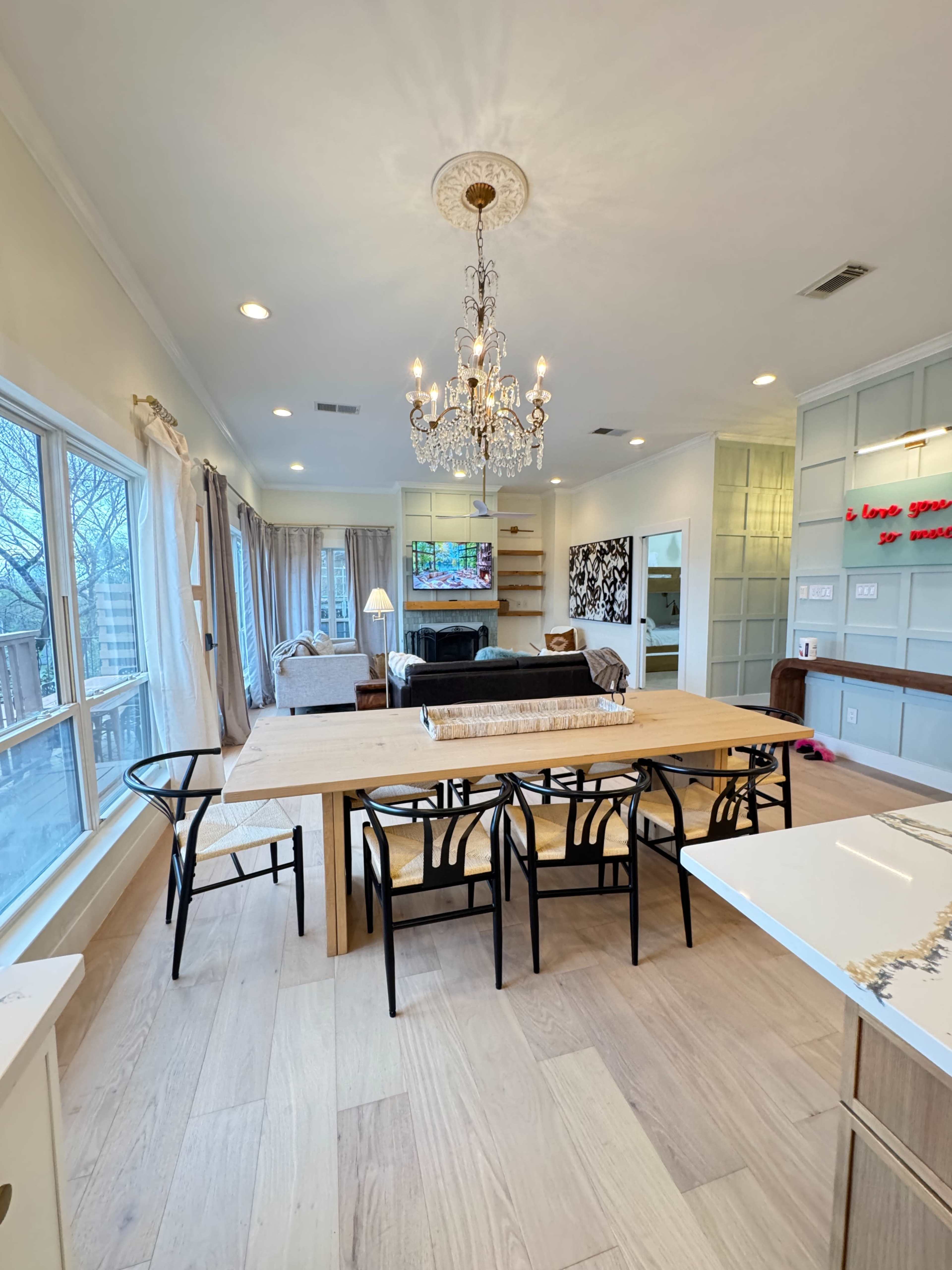 The image shows a modern dining area with a wooden table and black chairs, illuminated by a chandelier, featuring large windows and a living space in the background.