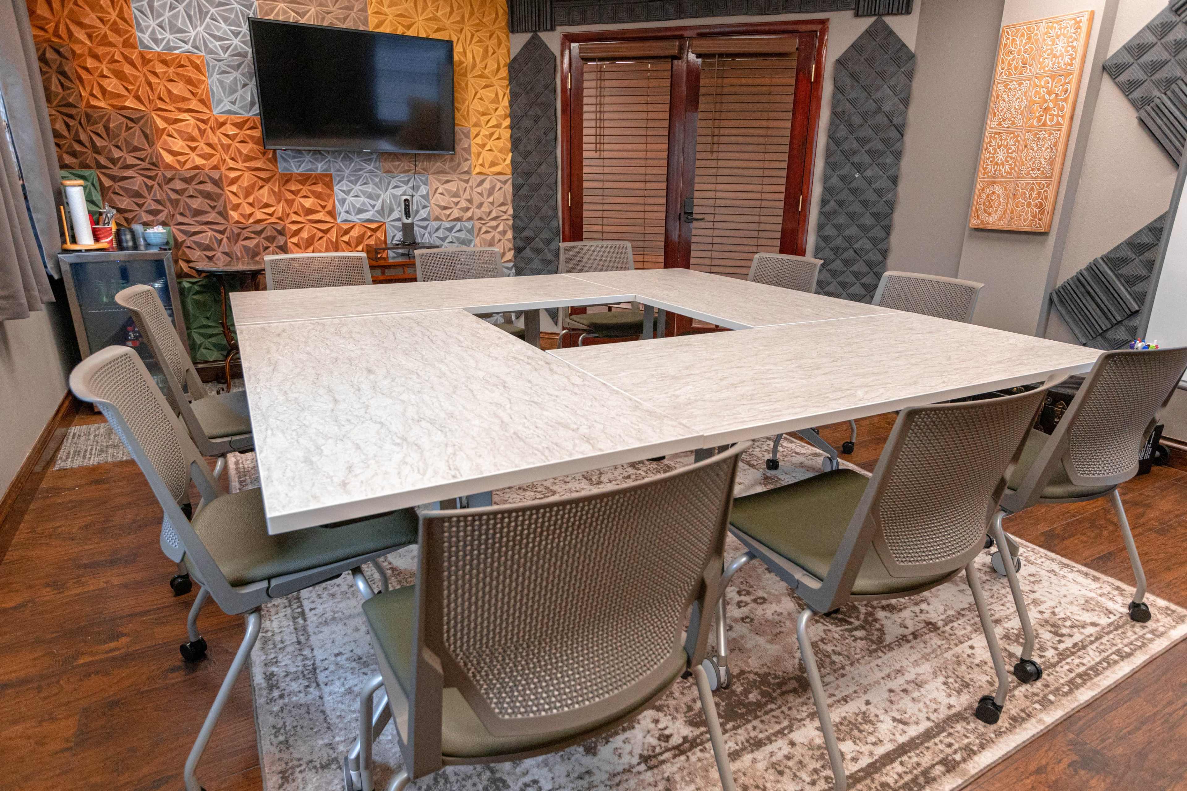 A large rectangular conference table is surrounded by several chairs in a room with textured wall panels and a television mounted on the wall.