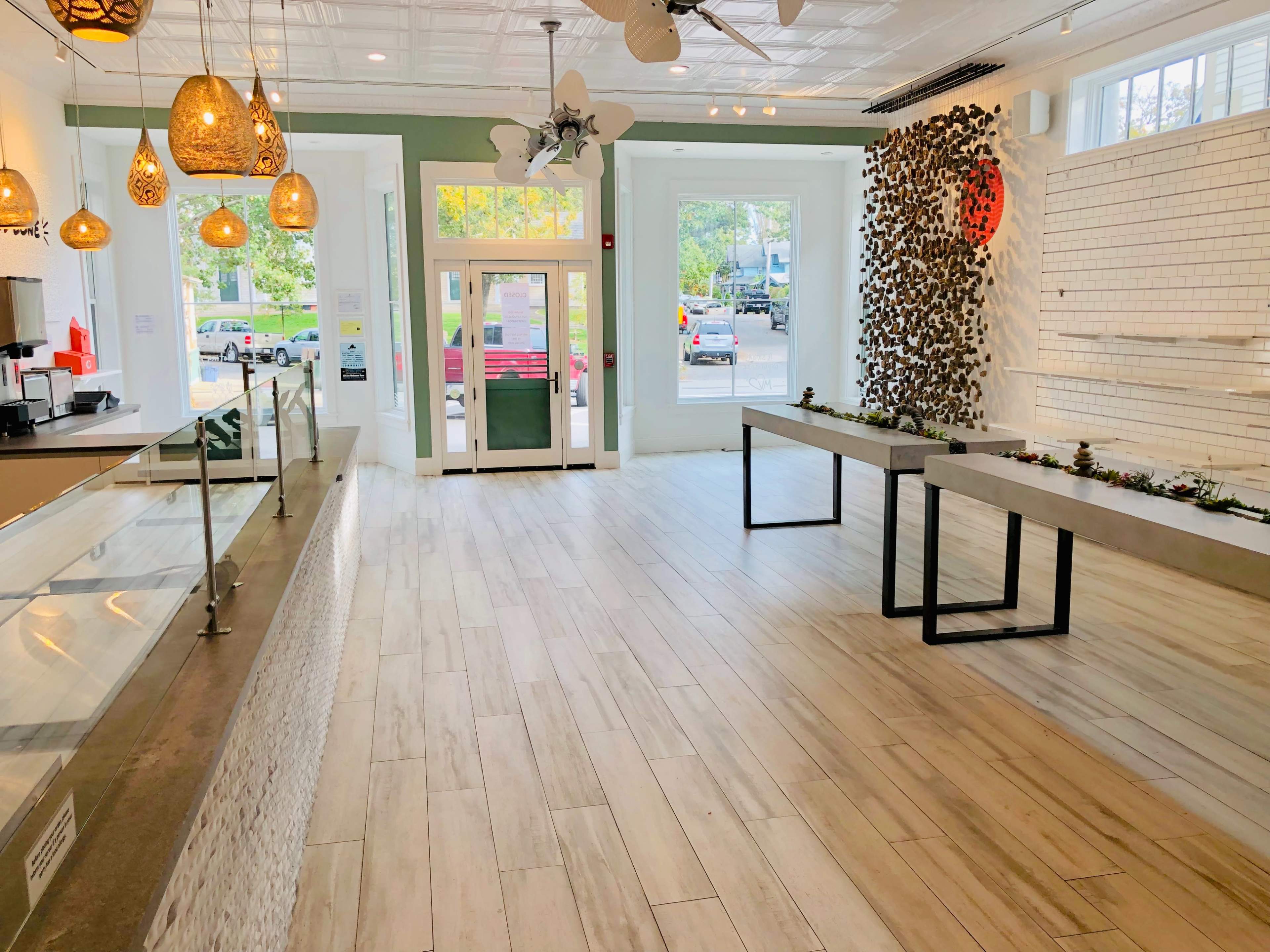 A modern interior of a cafe with light wooden floors, large windows, and minimalistic decor featuring hanging light fixtures and two metal tables.