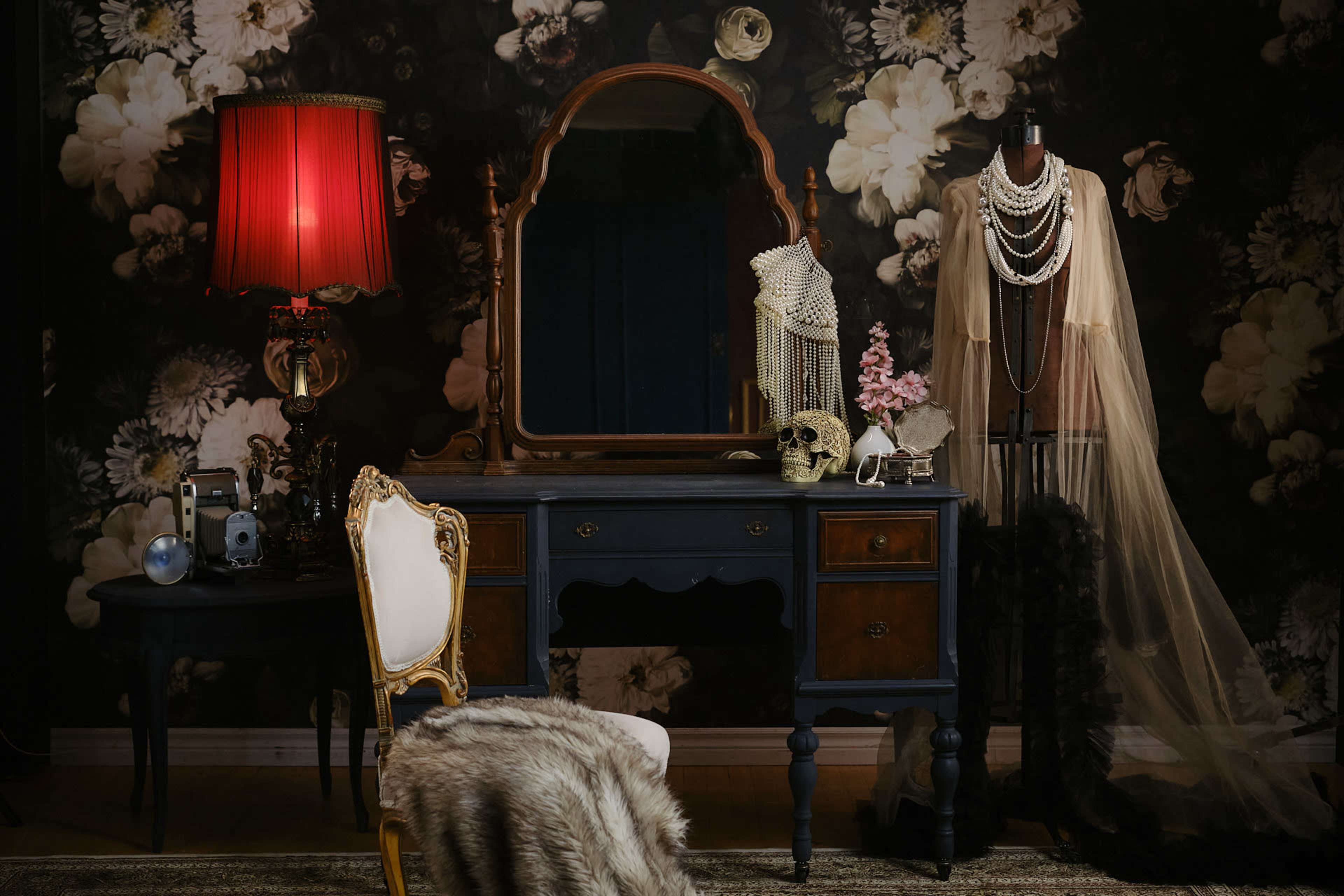 A vintage vanity table with a large mirror, a decorative chair, a red lamp, and a mannequin displaying elegant clothing and accessories, set against a floral wallpaper backdrop.