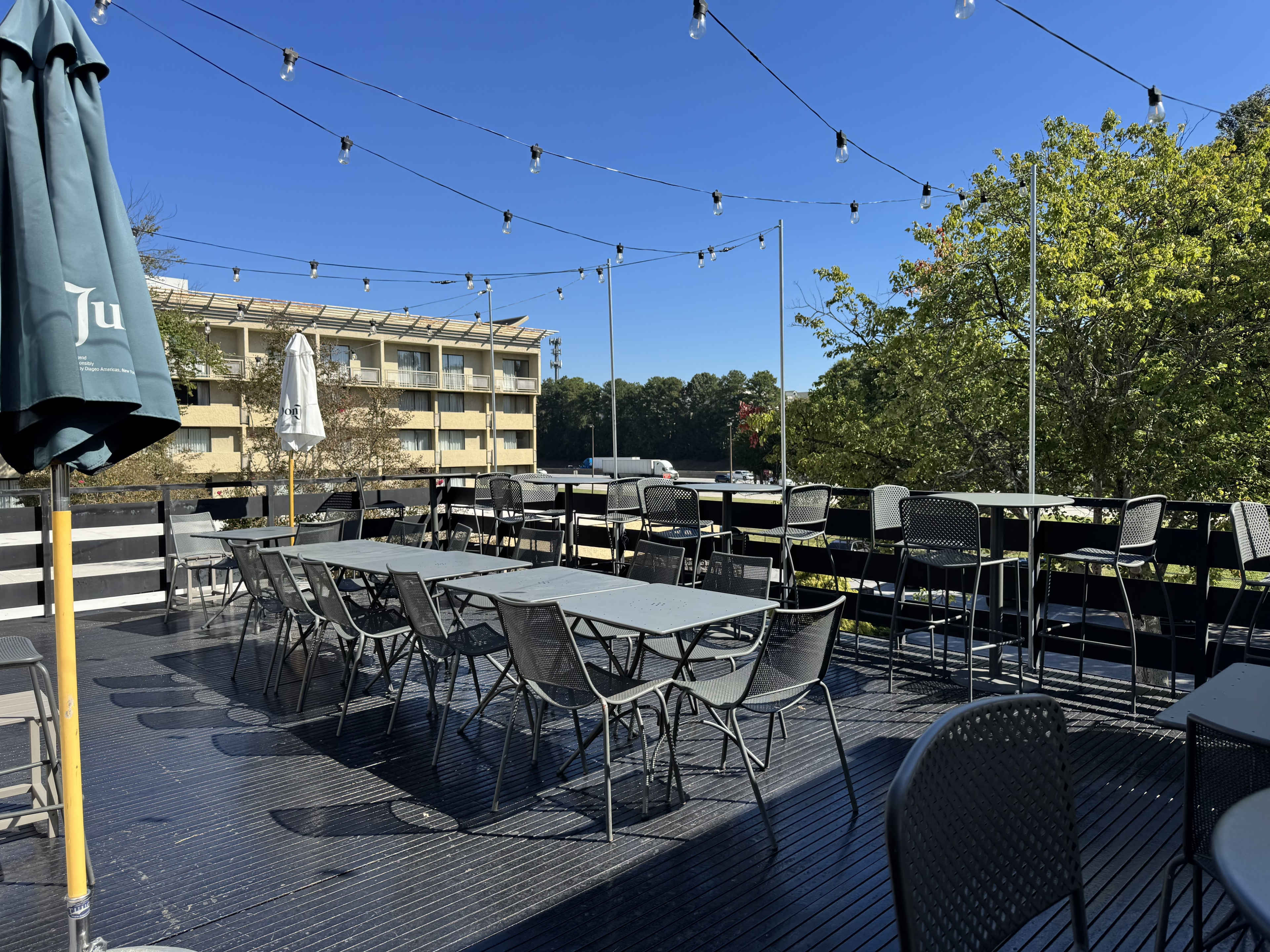 The image depicts an outdoor dining area with several tables and chairs positioned under string lights, adjacent to a building and surrounded by trees.