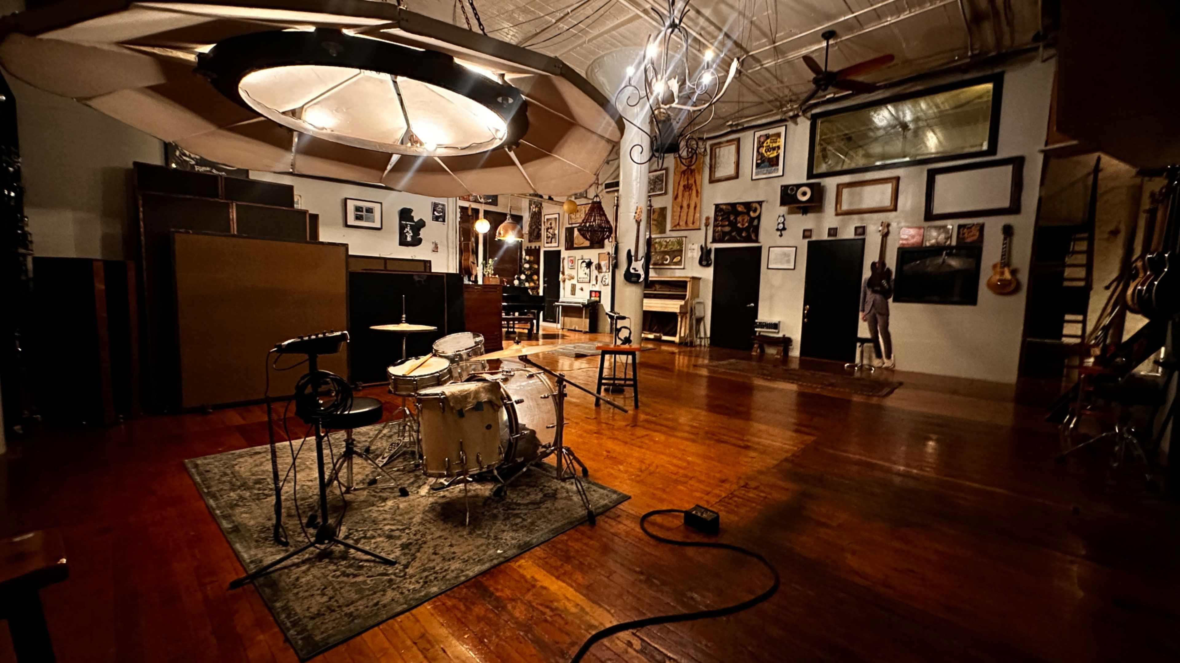 The image shows a room designed for music recording, featuring a drum set on a rug, wooden floors, and walls adorned with various framed artworks and musical instruments.