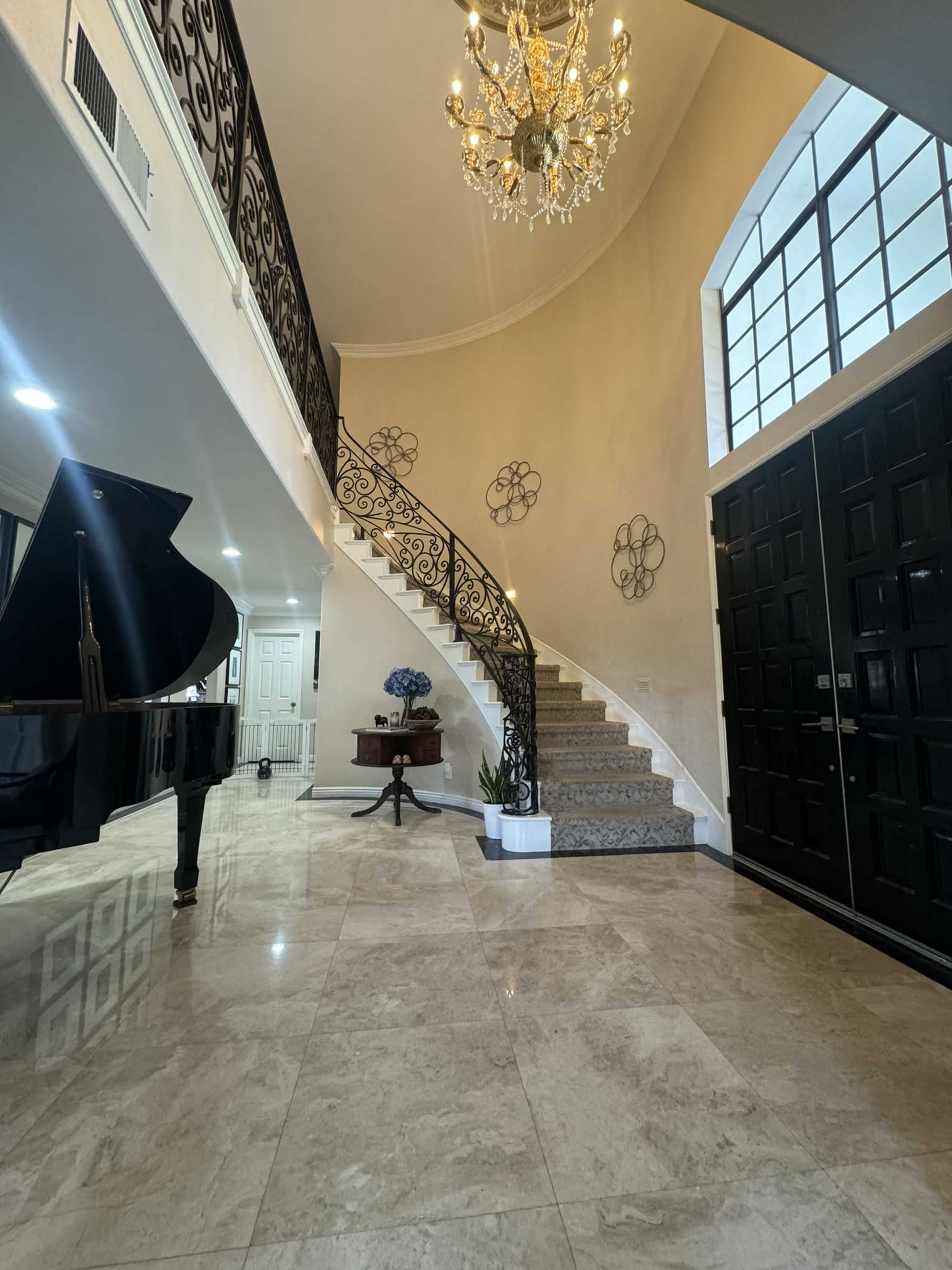 An elegant entryway featuring a grand staircase, a black piano, and a chandelier, with marble floors and decorative wall elements.