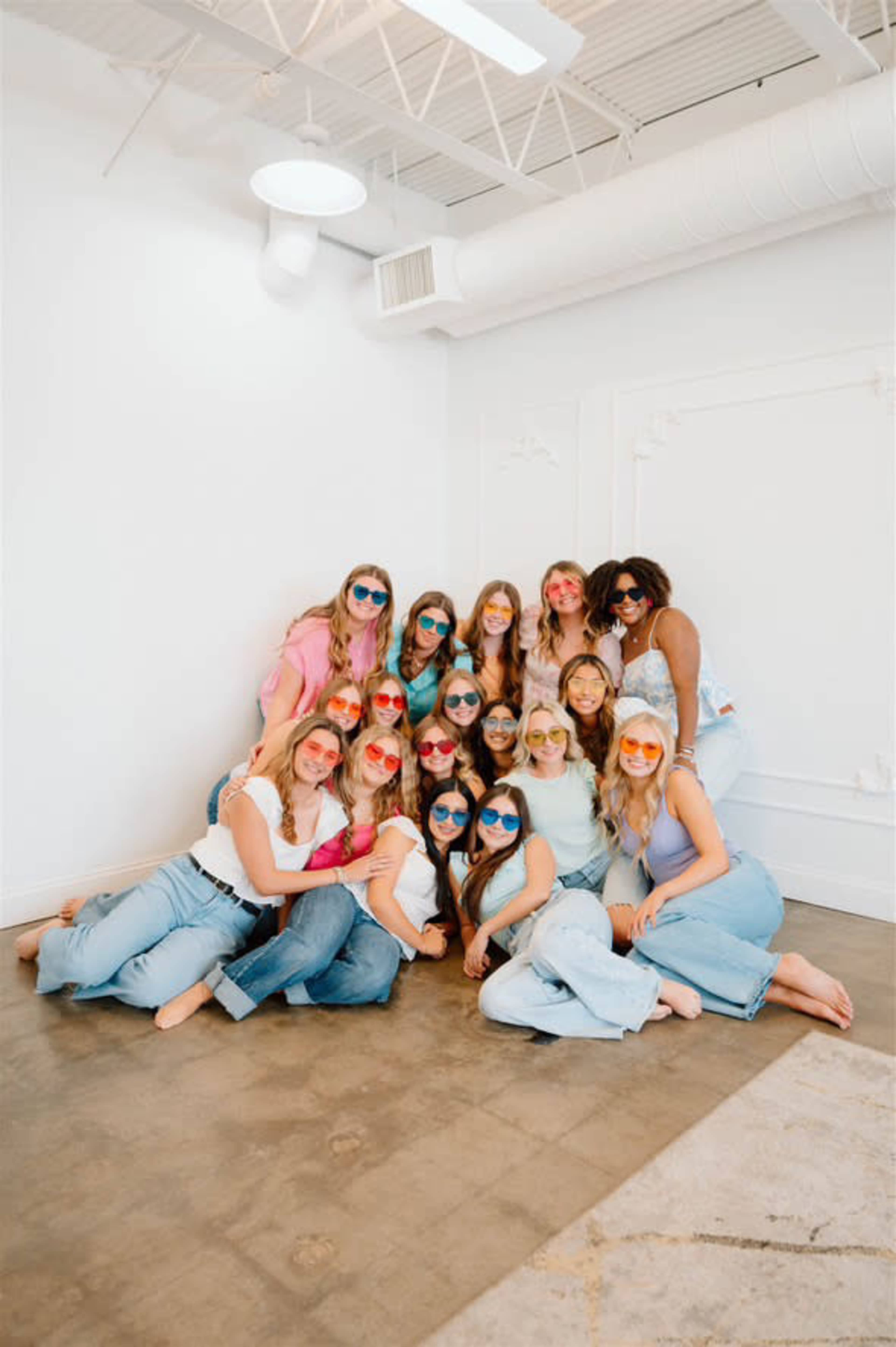A group of fifteen young women poses together while wearing colorful sunglasses in a bright, minimalistic indoor setting.