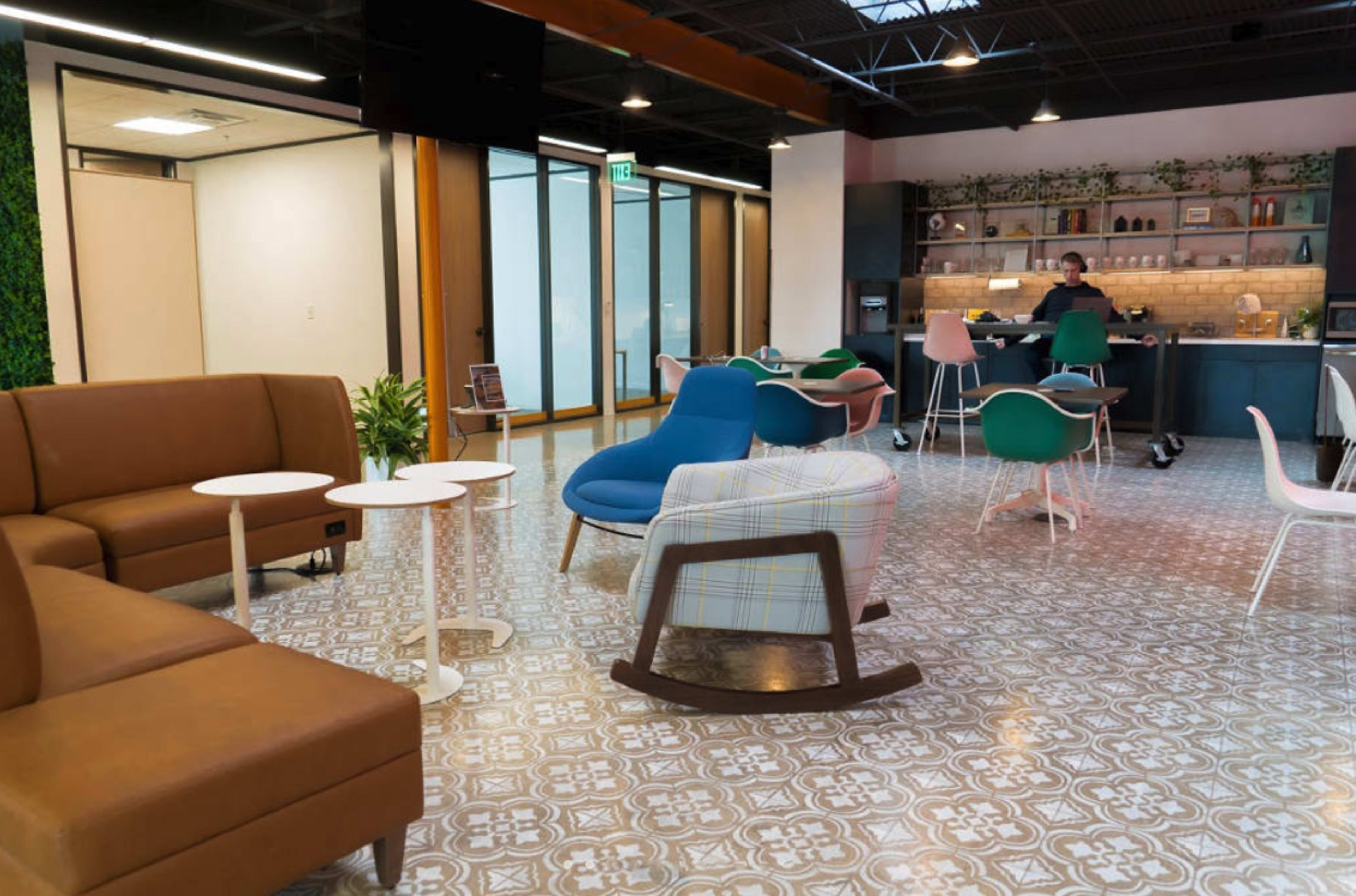 A modern lounge area with various seating options, decorative plants, and a kitchenette in the background.