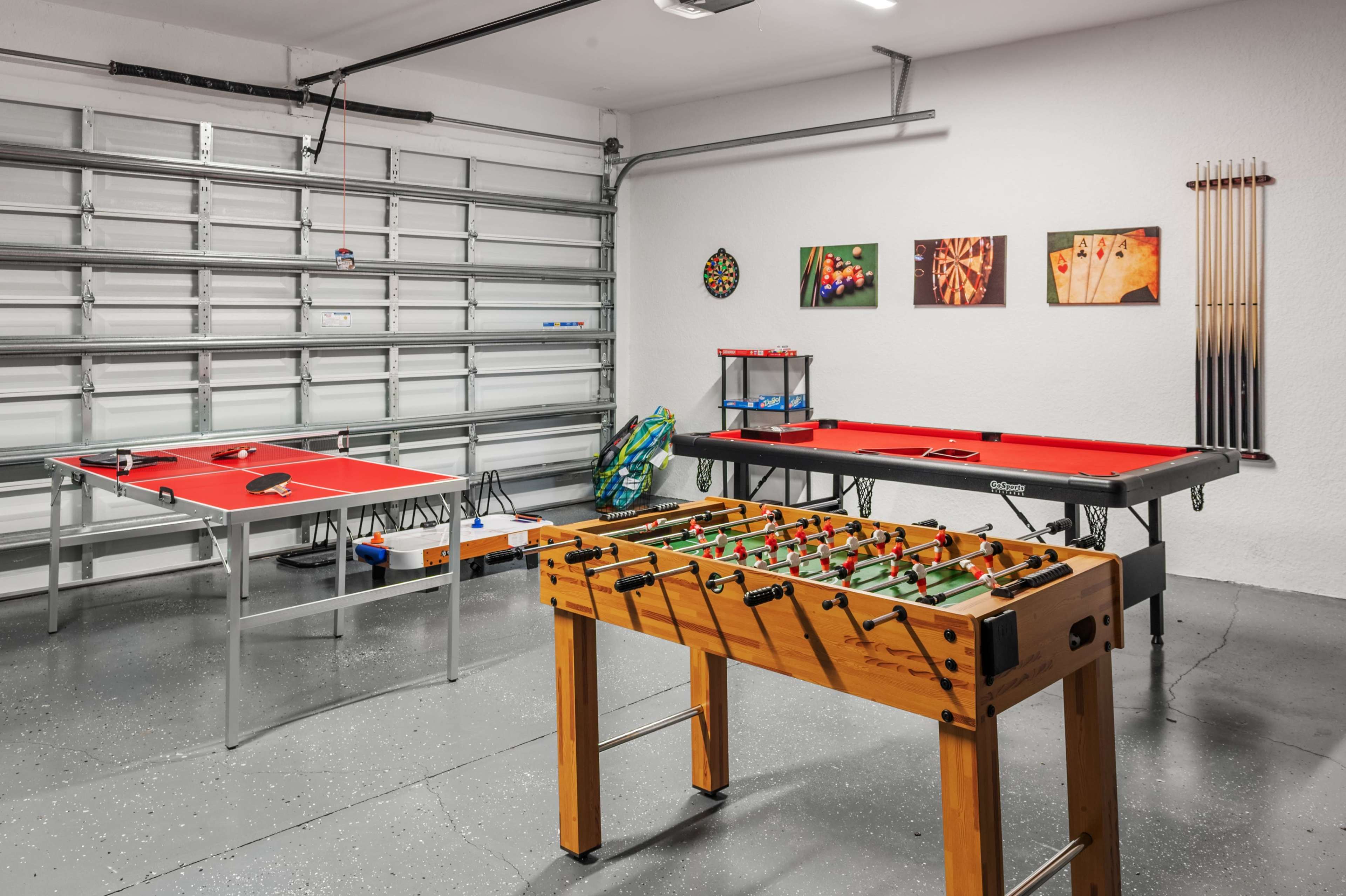 The scene features a garage game room with a foosball table, pool table, and air hockey table, along with wall art and a garage door in the background.