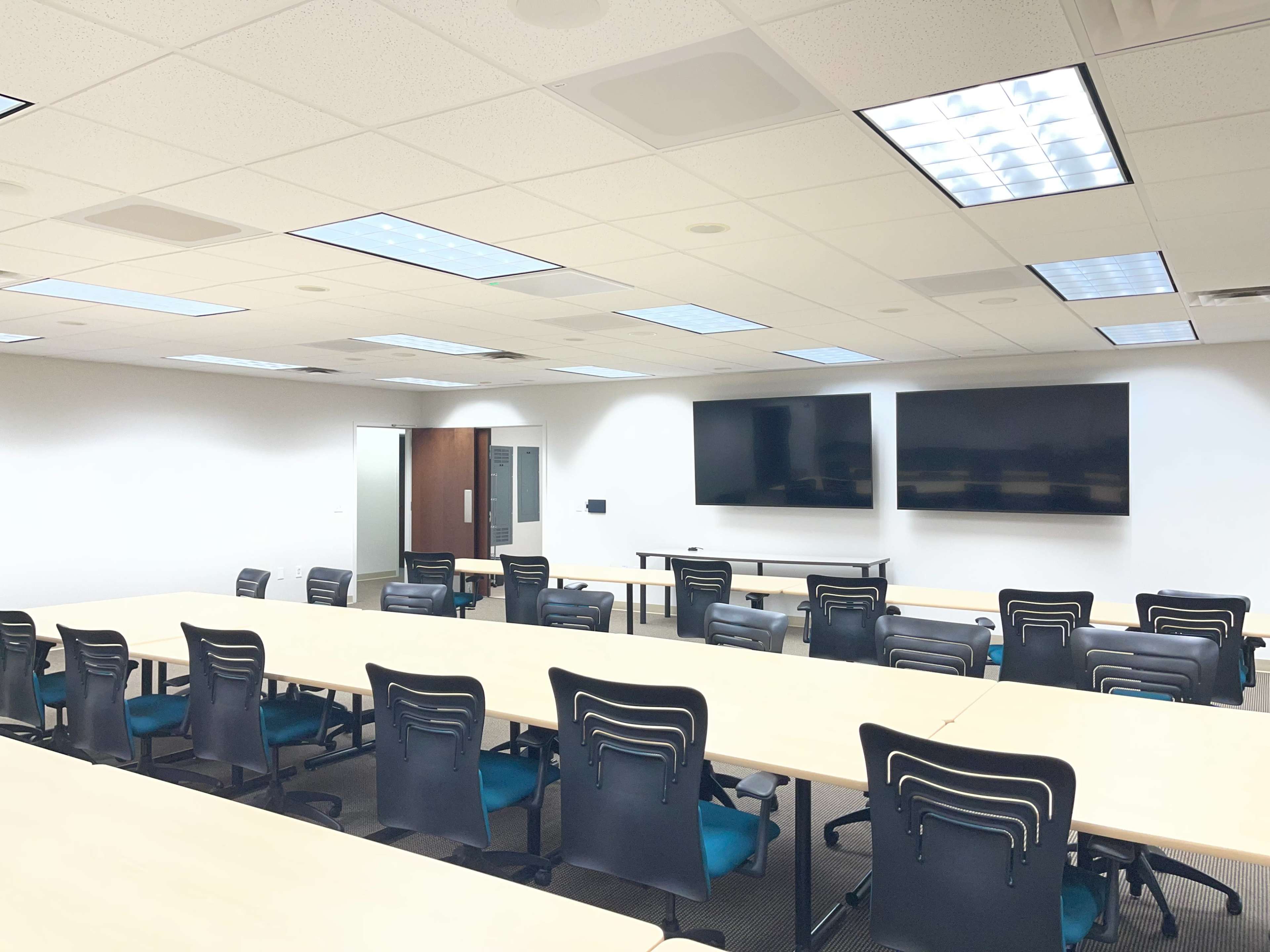 The image shows a large, empty conference room with multiple tables arranged in a U-shape and two large monitors mounted on the wall.
