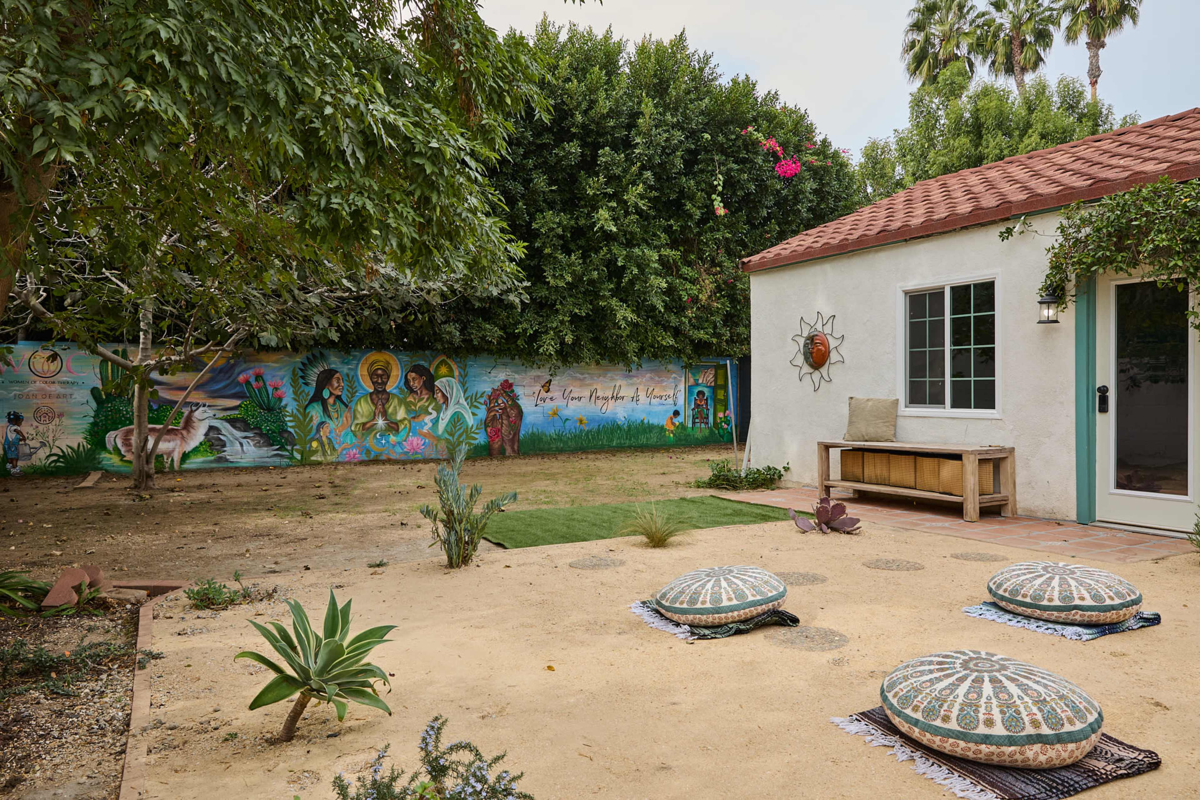 A serene outdoor space features three patterned poufs arranged on a sandy surface, with a mural depicting religious figures on a wall in the background.