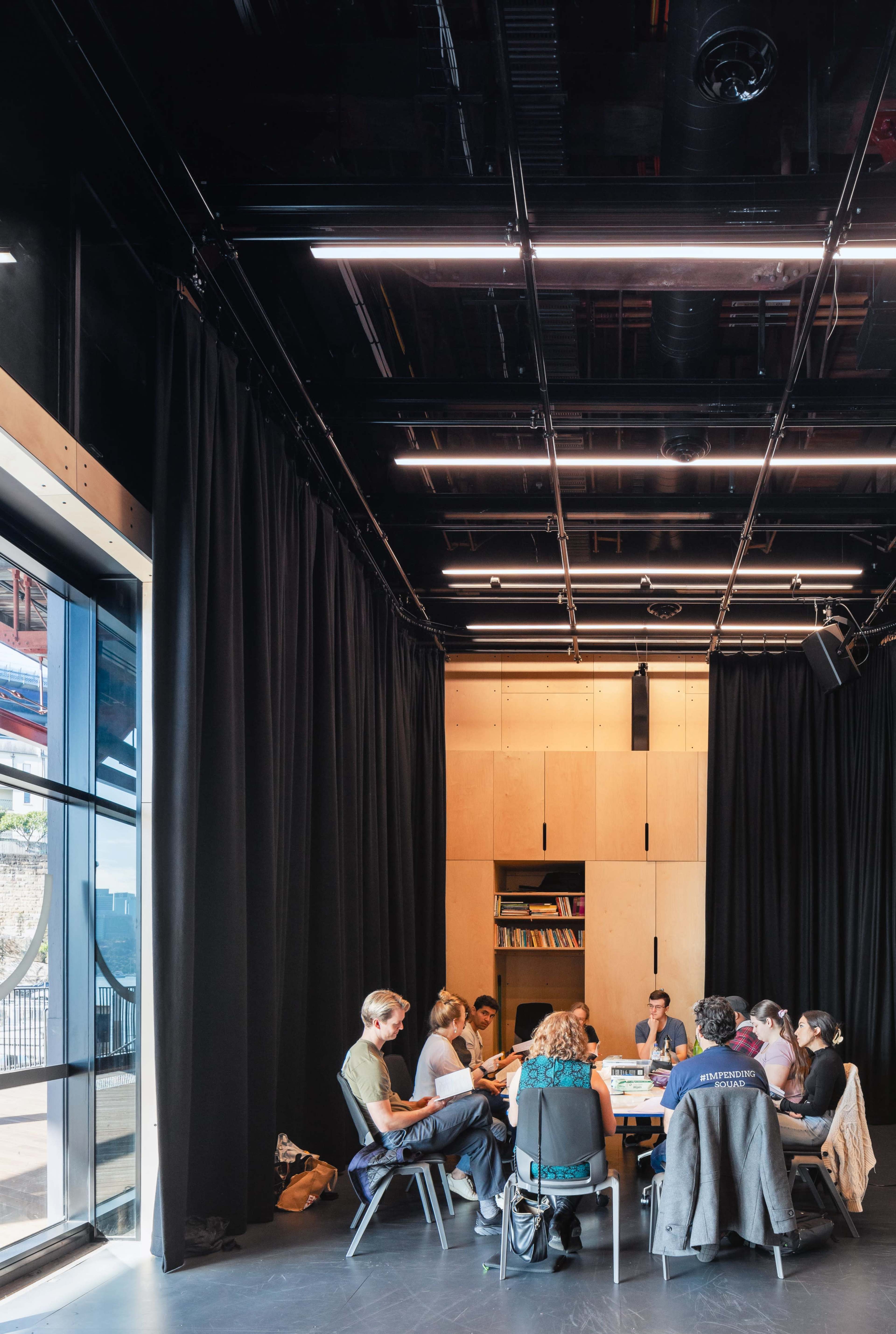 A group of people is gathered around a table in a modern, dimly-lit meeting room with large windows and black curtains.