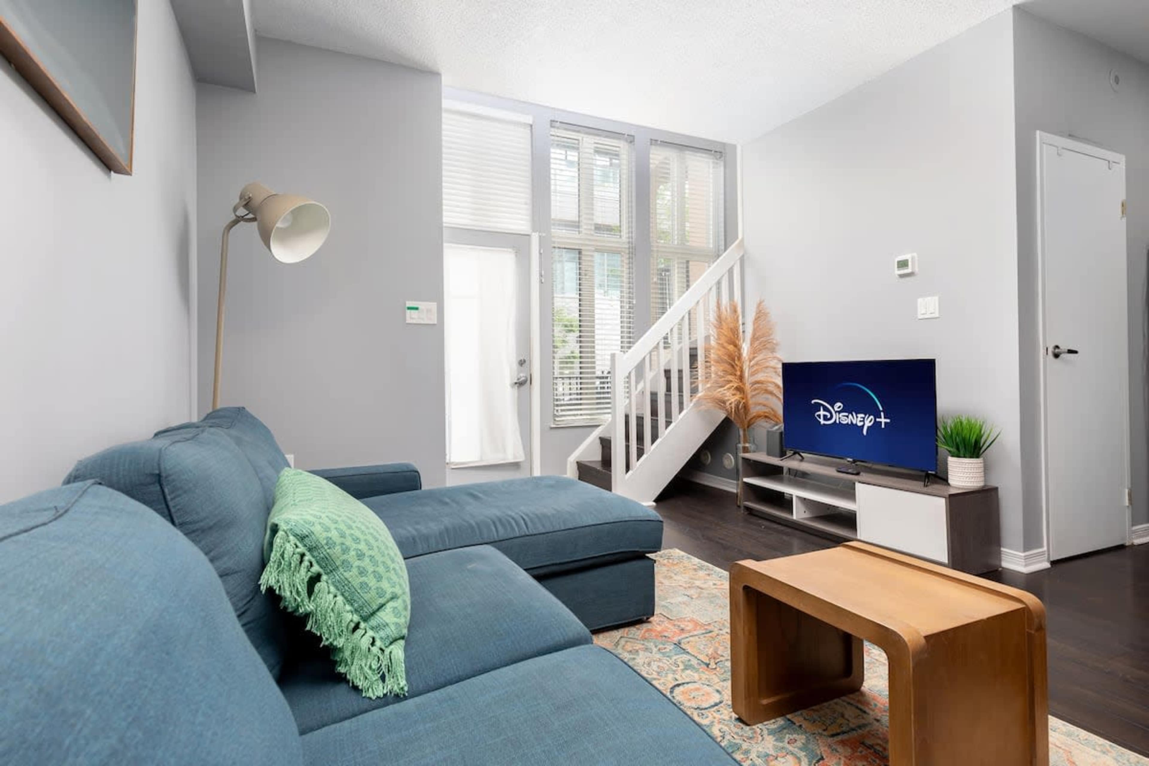 The image shows a modern living room with a teal sectional sofa, a wooden coffee table, and a television displaying the Disney+ logo, accompanied by large windows and a staircase in the background.
