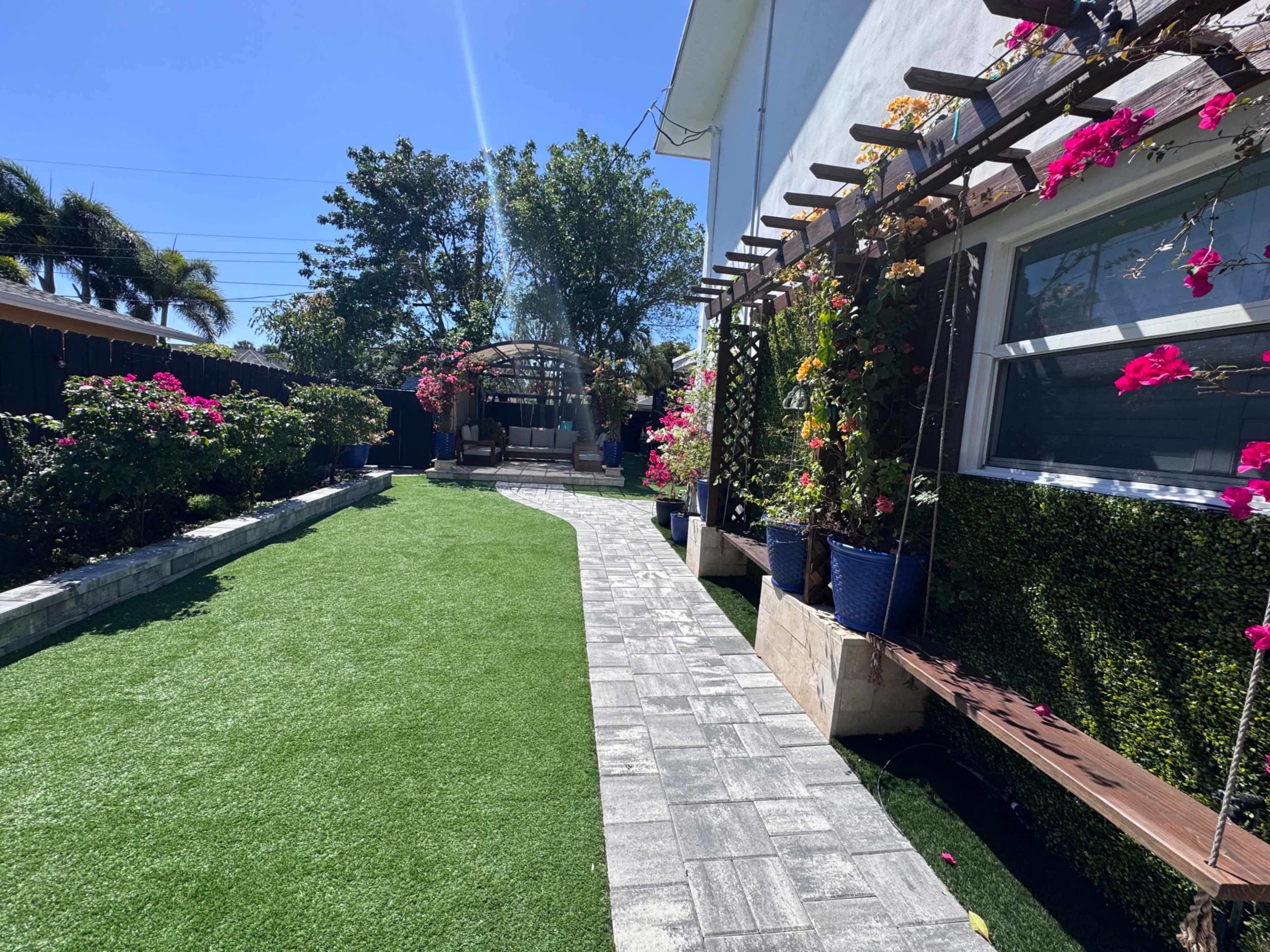 The image shows a landscaped backyard featuring a pathway lined with artificial grass, colorful flowers in pots, and a seating area under a pergola.