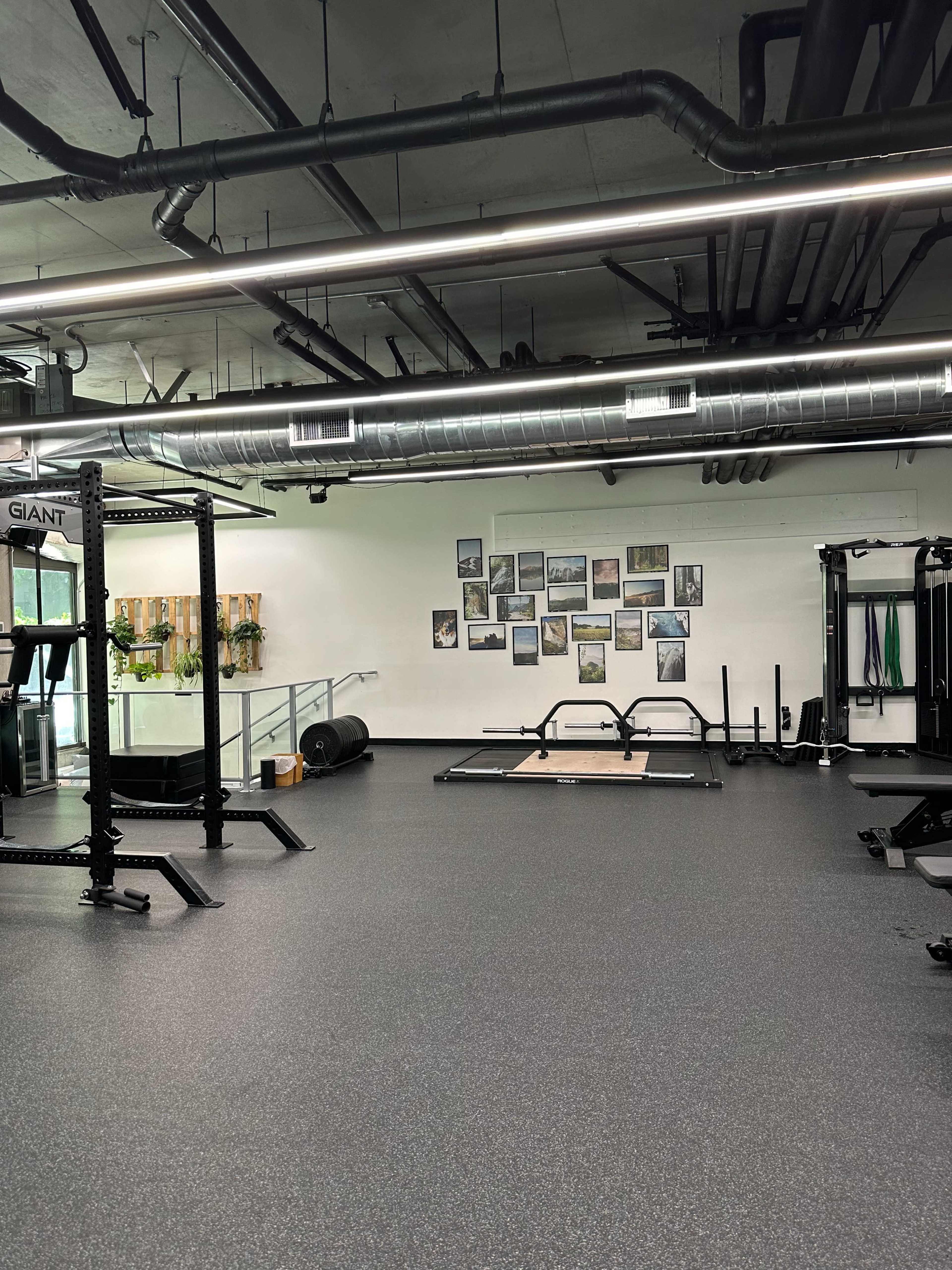 A spacious gym interior with workout equipment, including a power rack and benches, under industrial-style ceiling pipes and overhead lights.