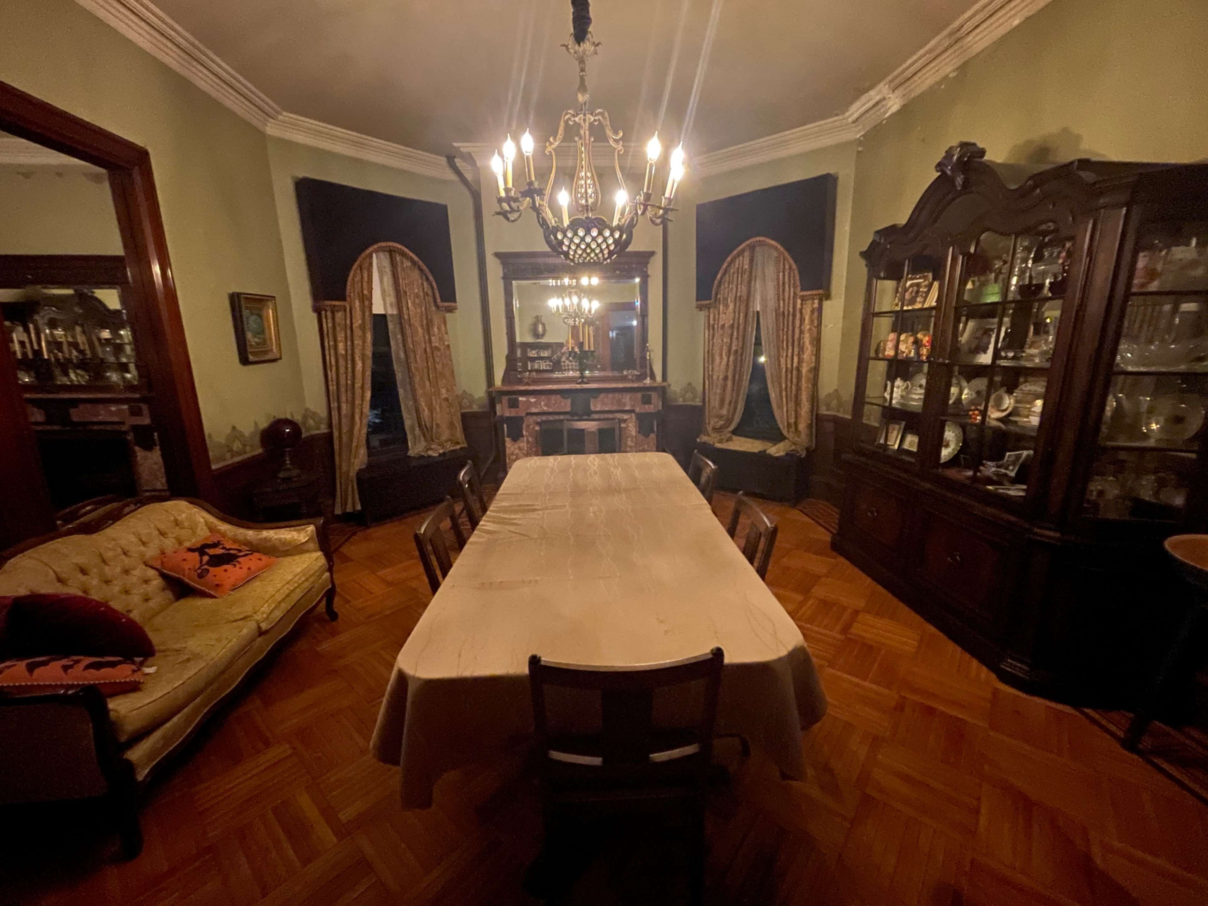 A dimly lit dining room with a large table surrounded by wooden chairs, flanked by a buffet and a china cabinet, all set against green walls and ornate curtains.
