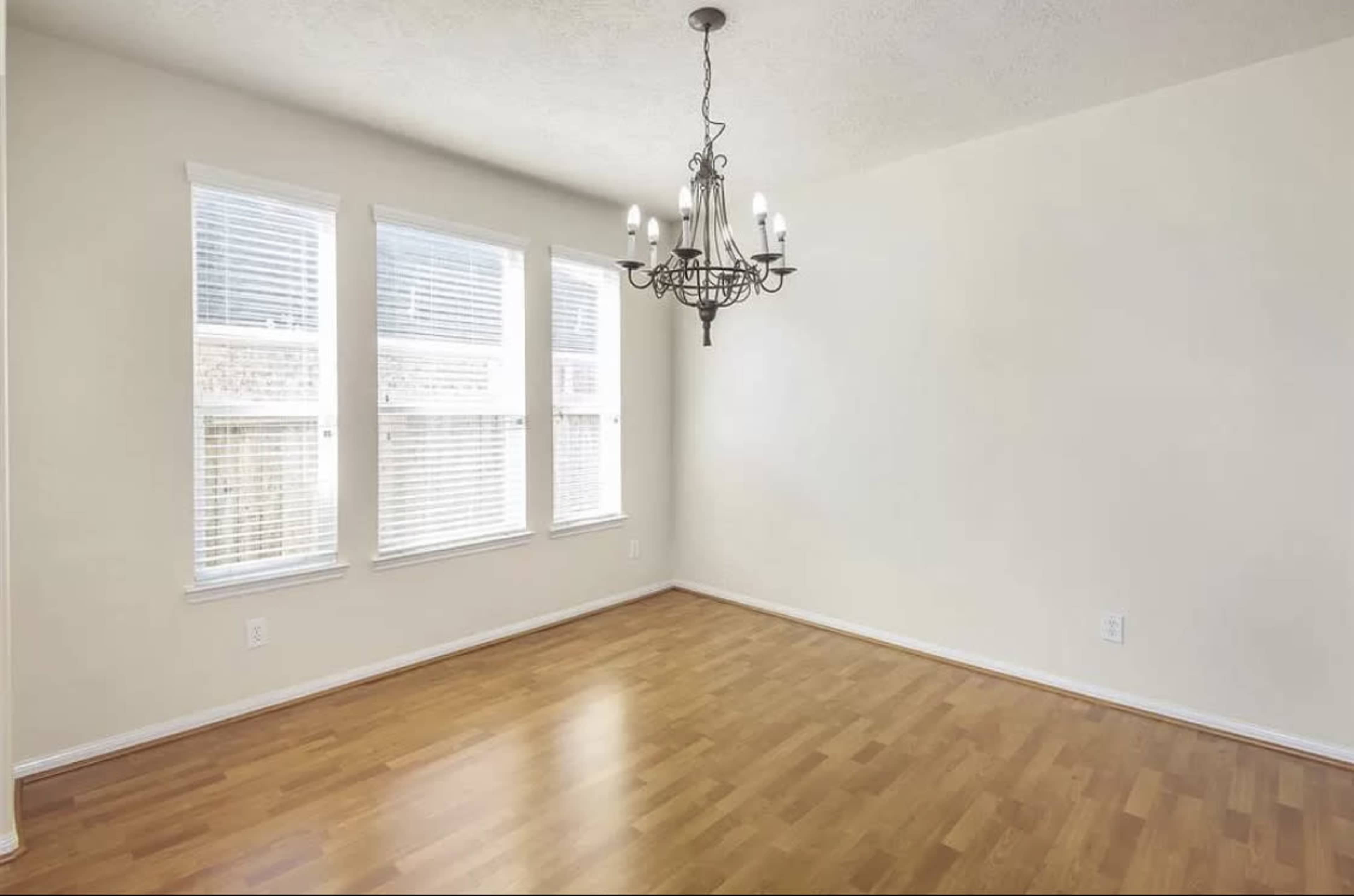 A simple, empty room with hardwood flooring, a chandelier, and three windows letting in natural light.